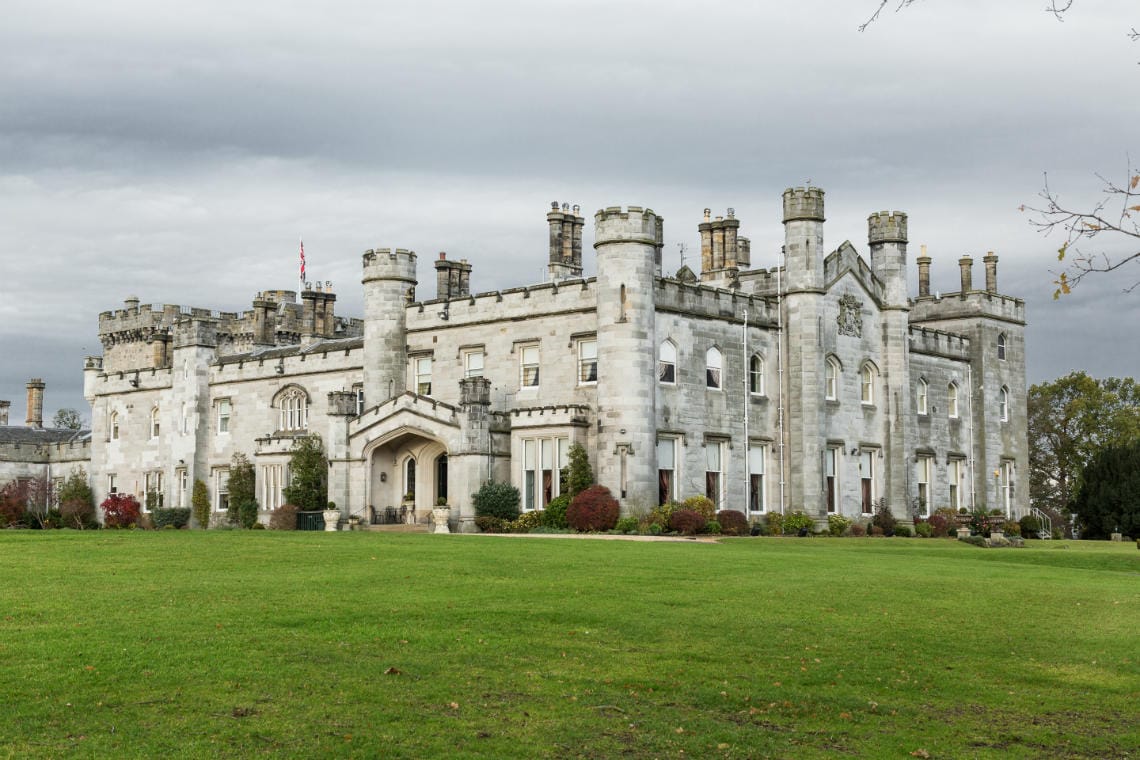 A large stone castle with multiple turrets and a flagpole, set on a well-maintained lawn under a cloudy sky.