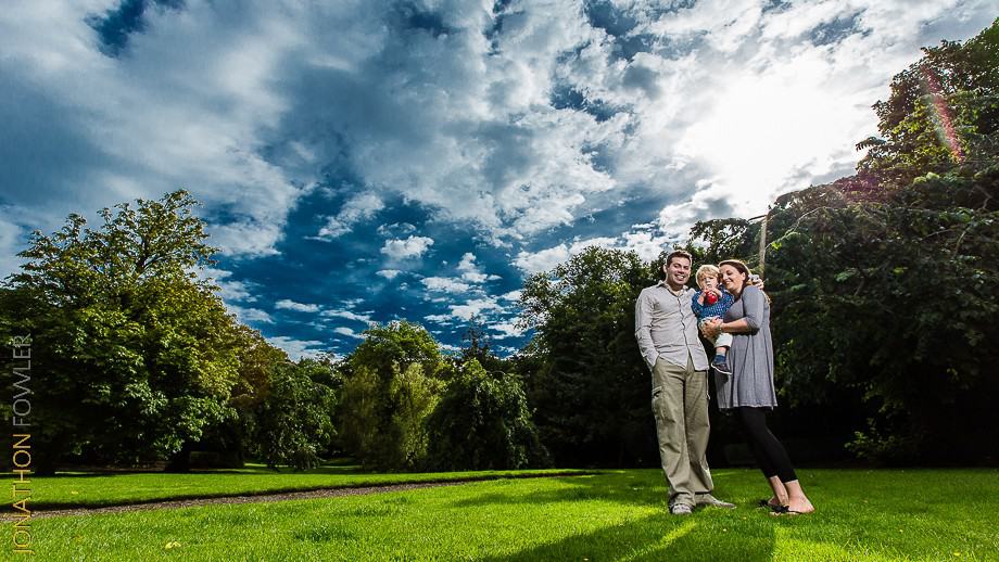 A man, a woman, and a child stand together on a grassy field with trees and a cloudy sky in the background.