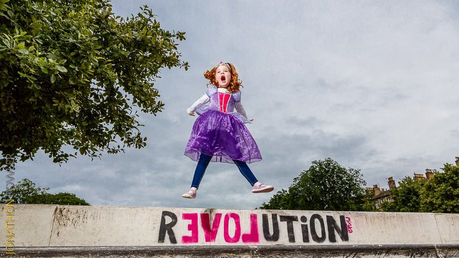 A child in a purple dress jumps on a concrete platform with "REVOLUTION" graffiti. Trees and buildings are in the background.