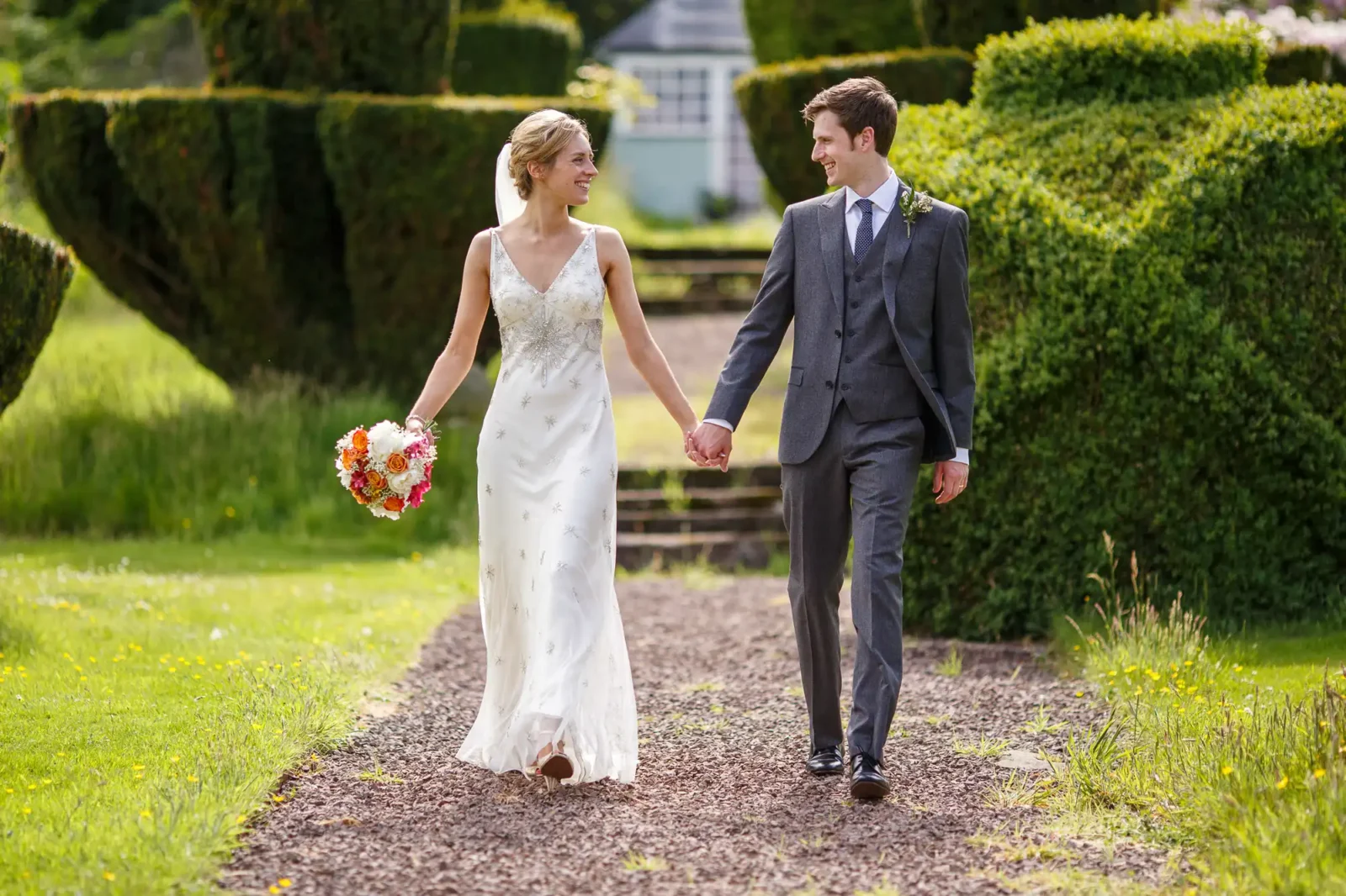 Fingask Castle wedding photos. A bride in a white dress and a groom in a grey suit walk hand in hand on a garden path, smiling at each other. The bride holds a bouquet of flowers.