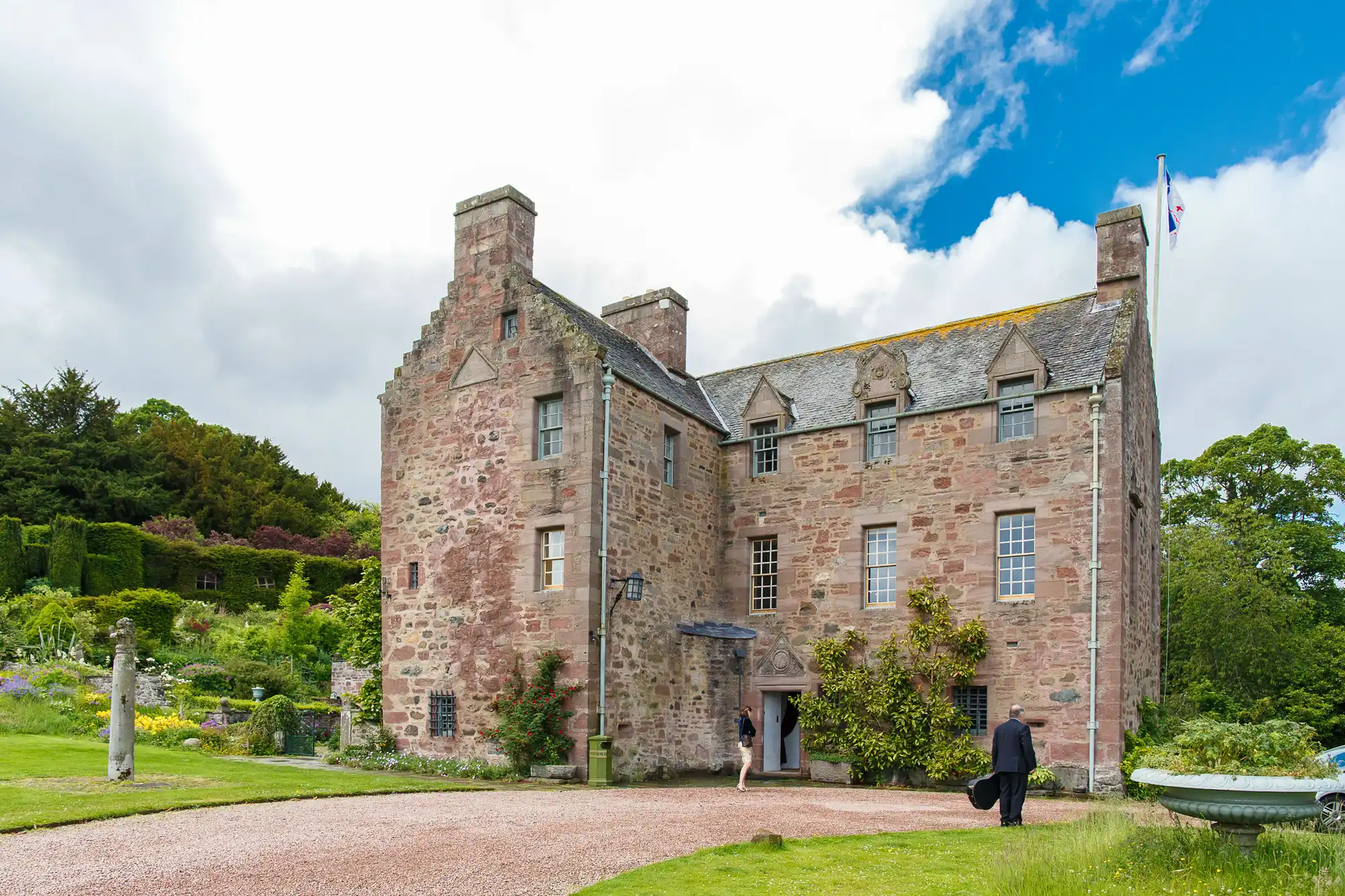 A person walks toward an old, stone castle with a scottish flag on a sunny day, surrounded by lush greenery and a vibrant garden.