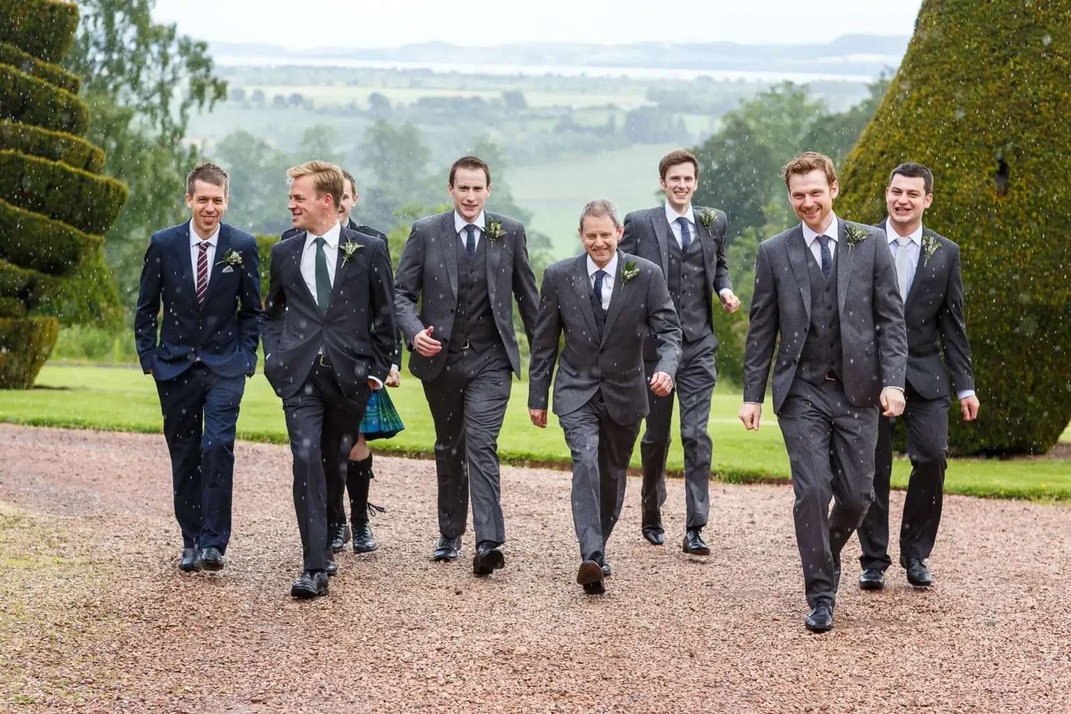 Wedding photography of seven groomsmen in suits walk outside on a gravel path, with a grassy landscape and overcast sky in the background. The two leftmost individuals wear ties with stripes, while the others have solid-coloured ties at Fingask Castle, Perth and Kinross.