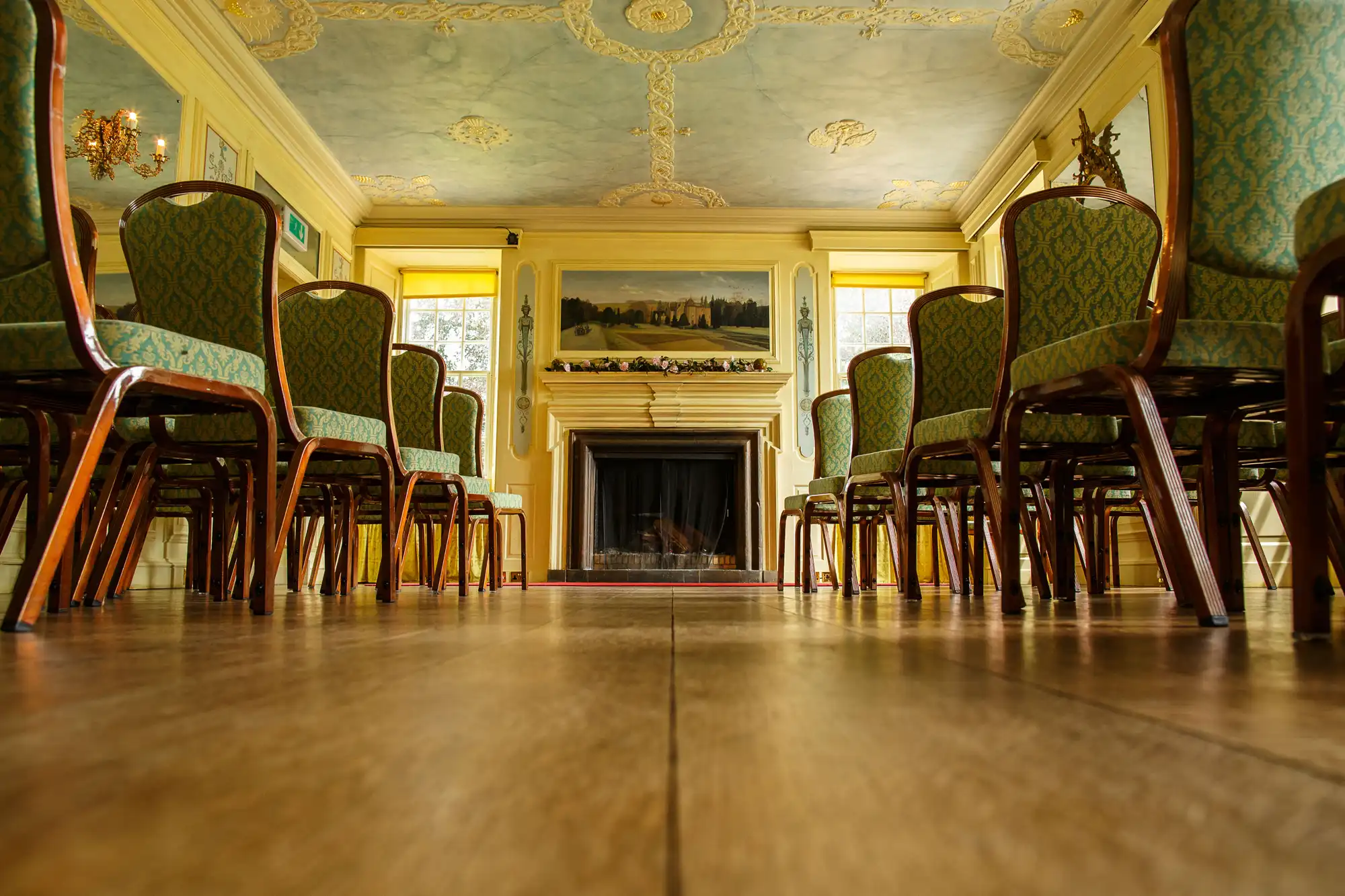 Low angle view of an ornate room with green upholstered chairs, polished wooden floor, and a fireplace under a classical painting.