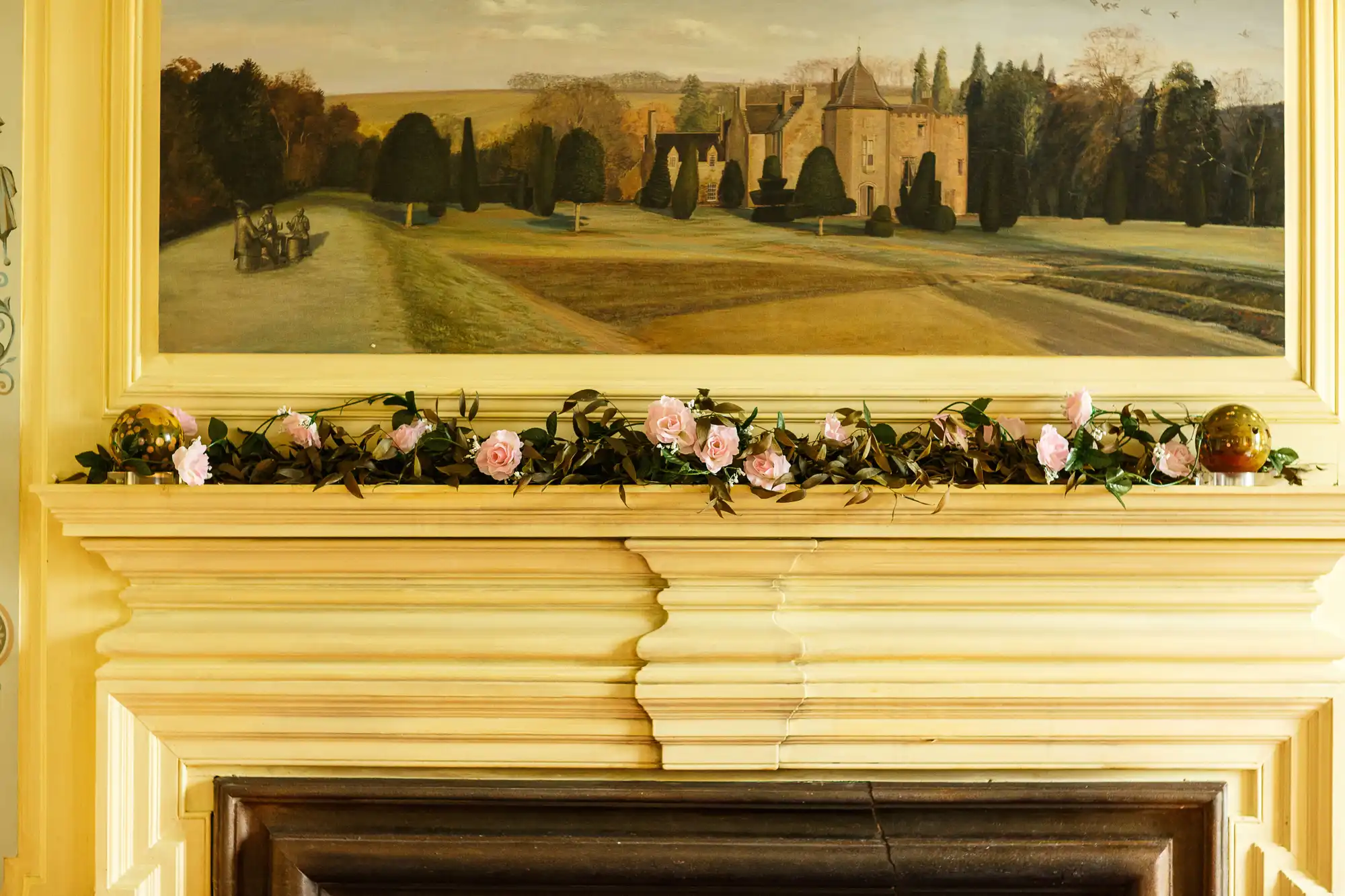 A painting of a castle and landscape hangs above a fireplace, adorned with a floral garland and decorative globes.