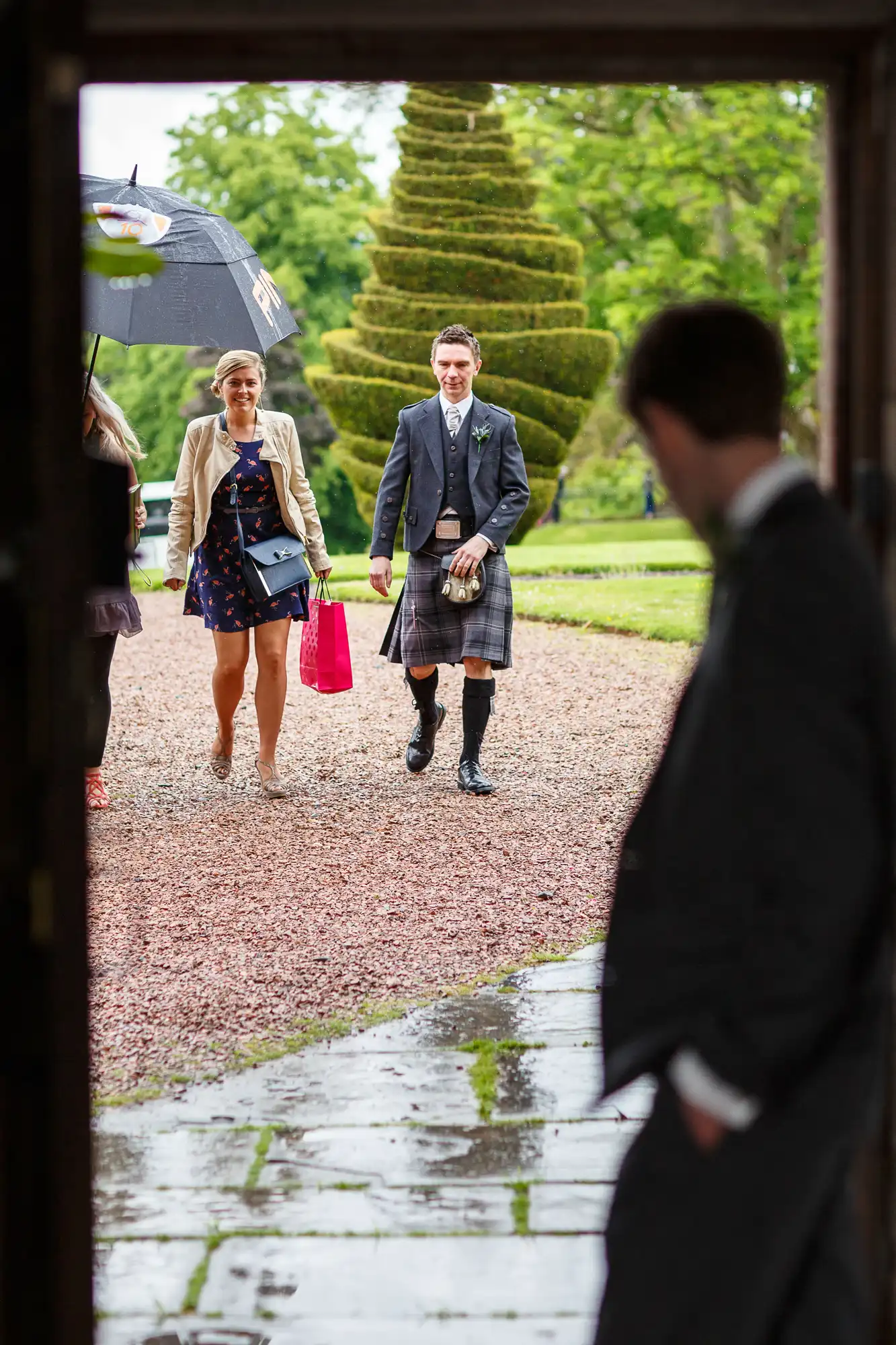 A couple dressed in formal attire walks towards a building under an umbrella on a rainy day, with a man's silhouette in the foreground.