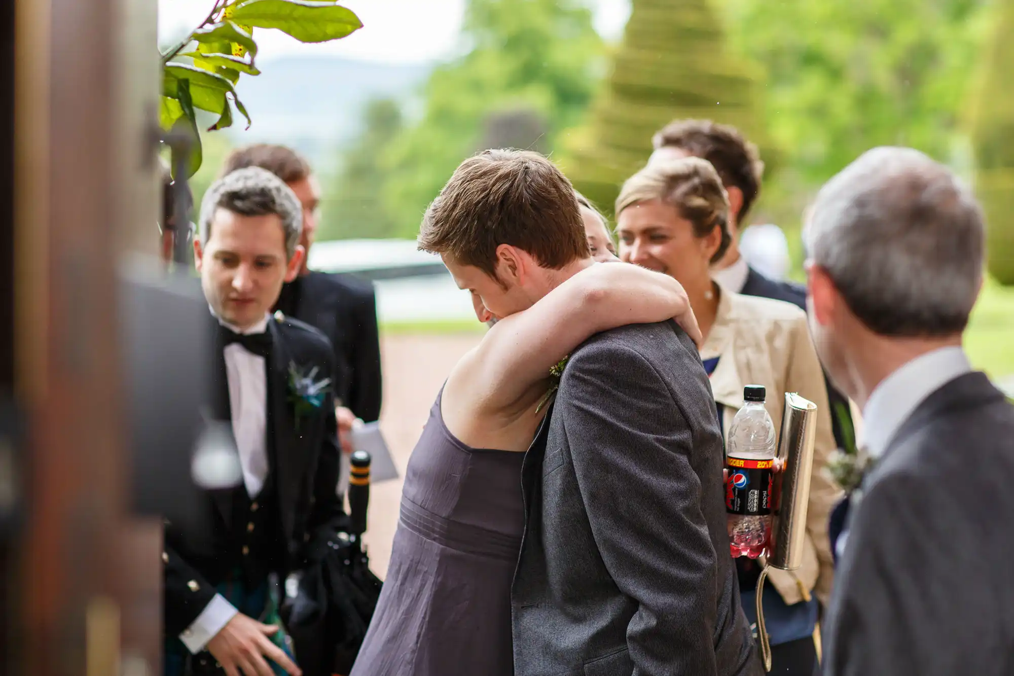 A man in a suit hugs a woman in a gray dress at an outdoor gathering, with other attendees visible in the background.