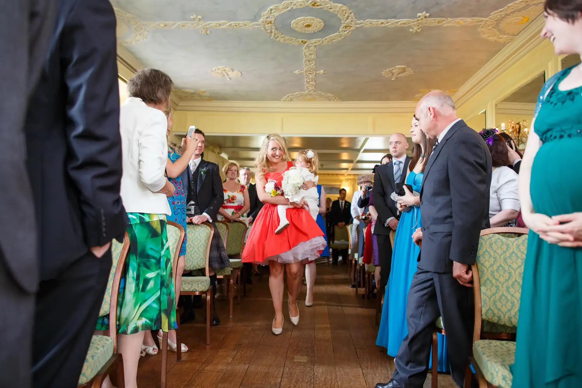 Bride in a white and red dress walking down the aisle, guests watching and taking photos in an ornate room.