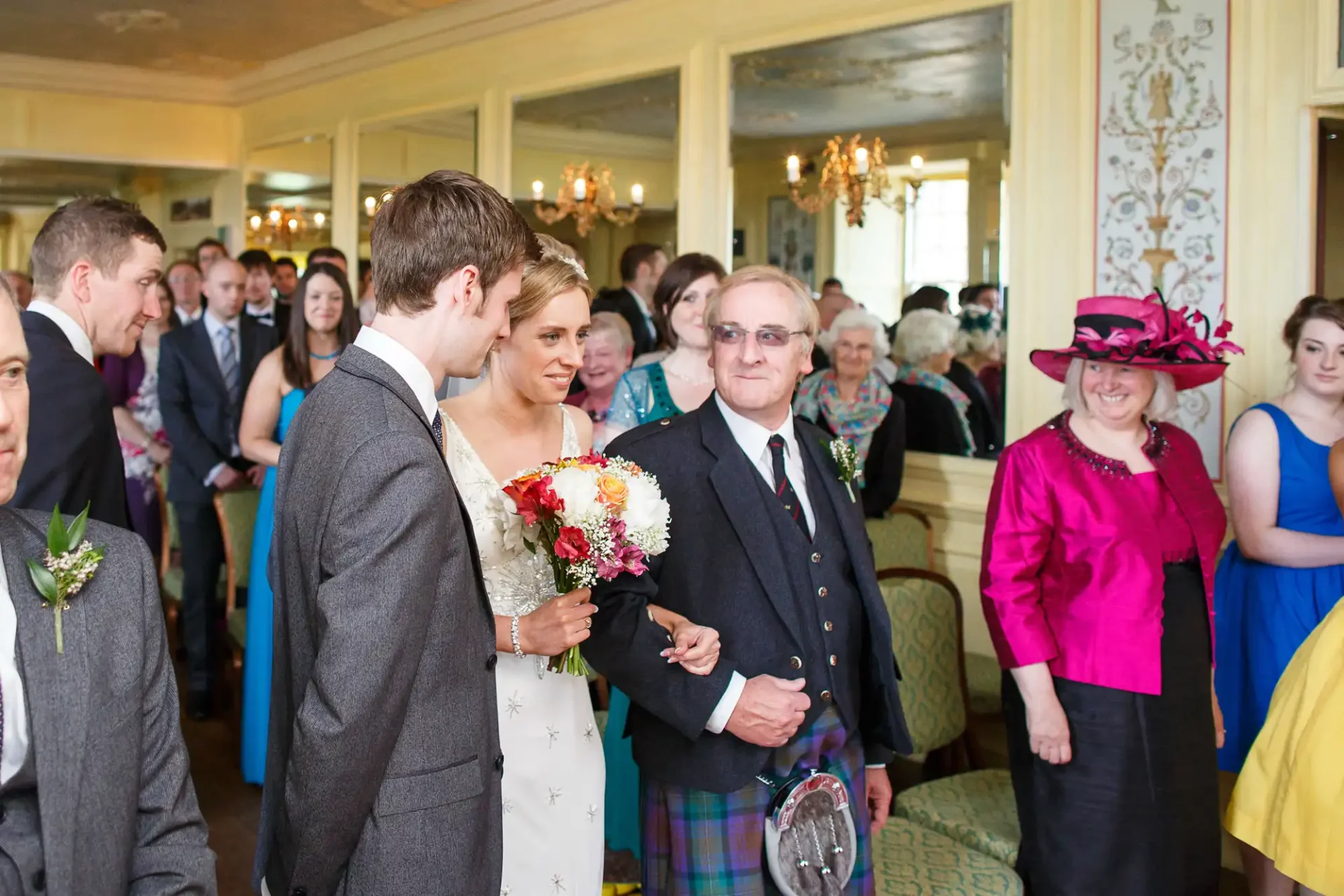 A bride and groom walk down the aisle hand in hand, surrounded by smiling guests in formal attire at a wedding ceremony.