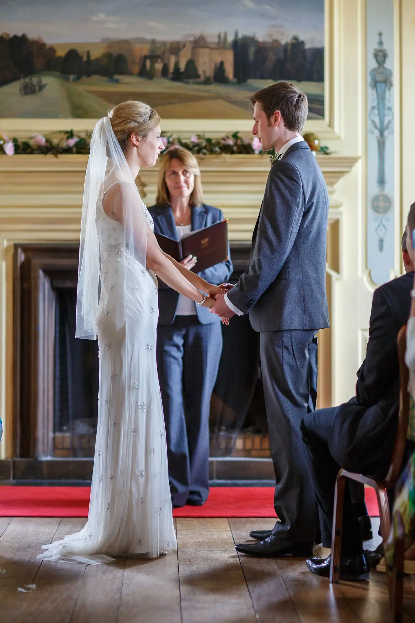 A bride and groom holding hands during wedding vows, with an officiant and guests in a room decorated elegantly.