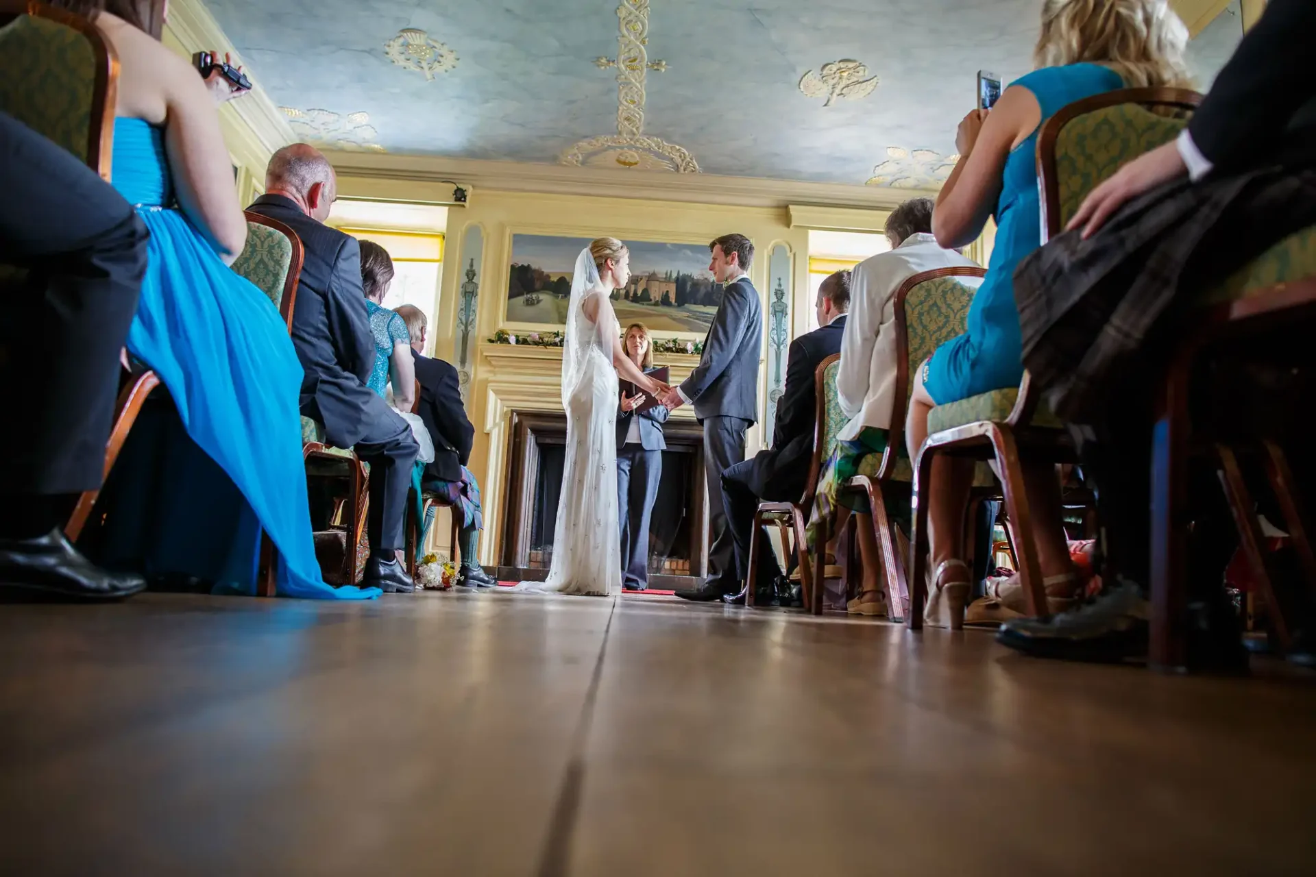 A bride and groom exchange vows in a grand room with ornate ceilings, surrounded by seated guests.