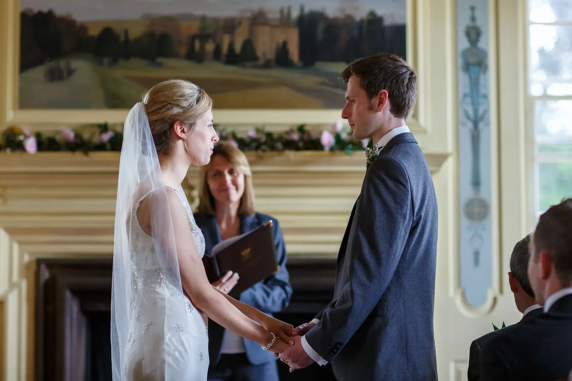 A bride and groom holding hands, exchanging vows in a formal wedding ceremony indoors, with a sunlit landscape visible through the window.