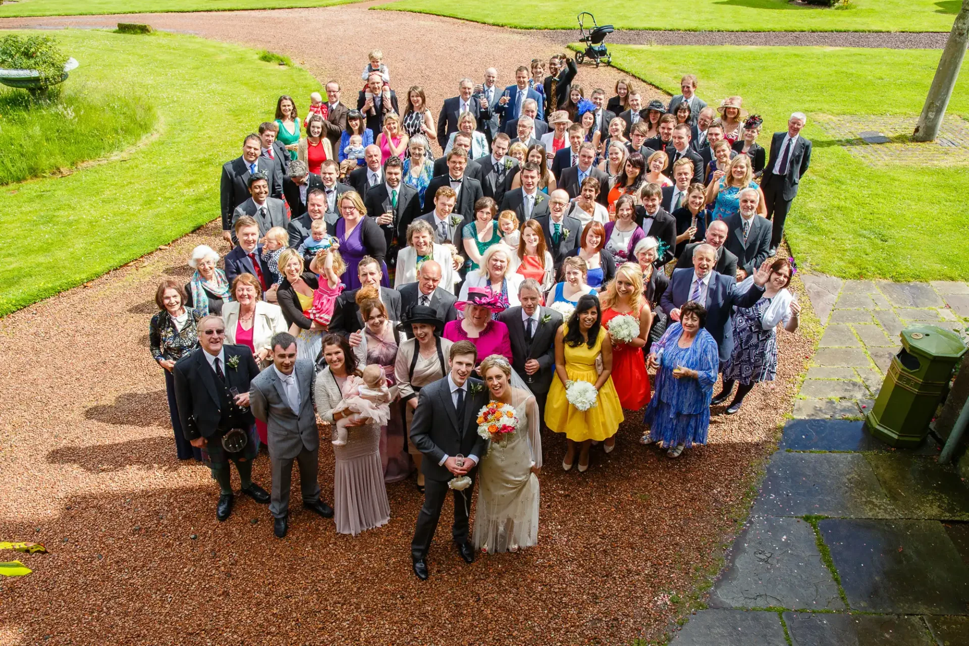 A large group of wedding guests posing with the bride and groom outdoors on a sunny day, some waving at the camera.