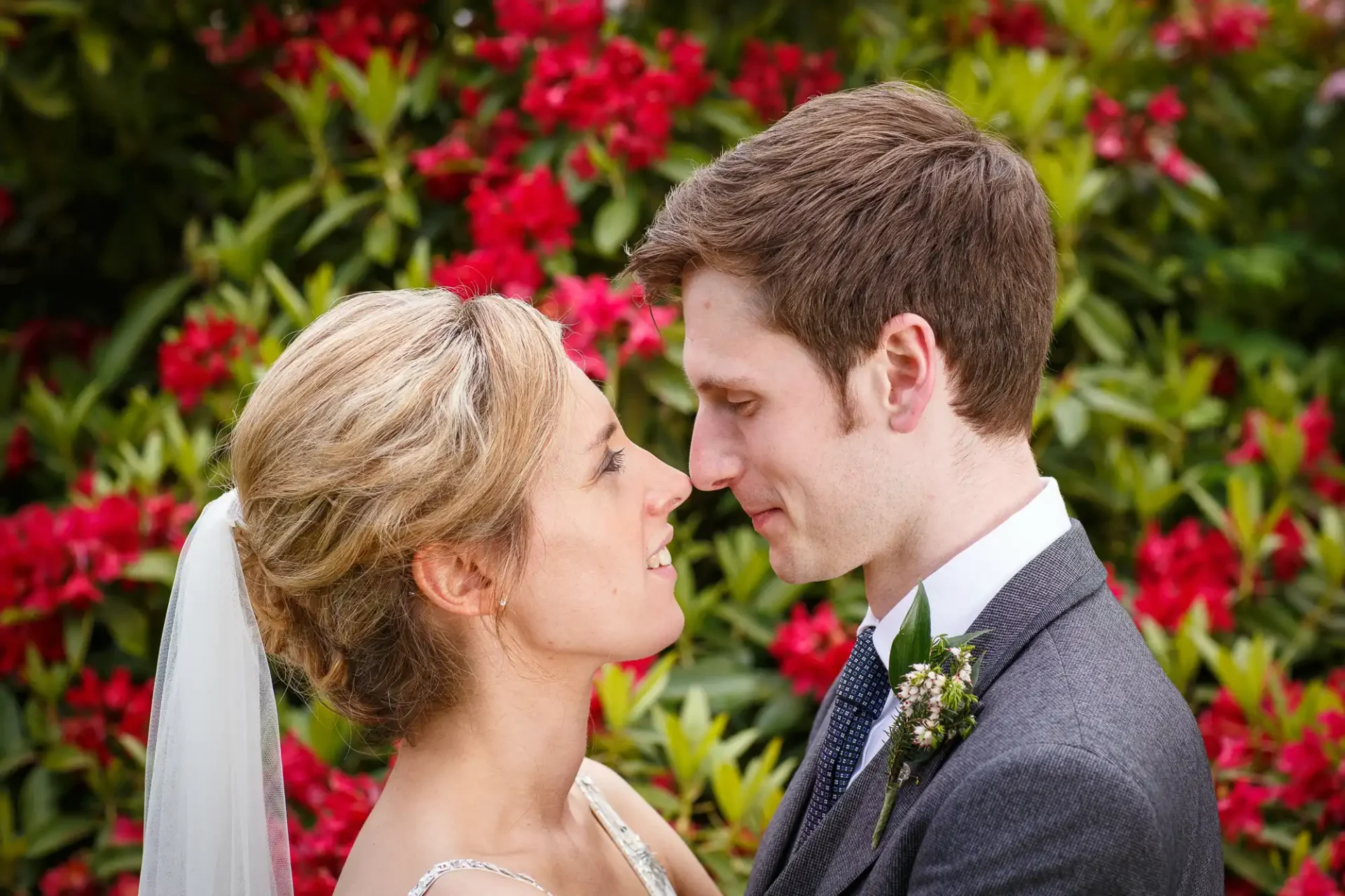 A bride and groom smile at each other, the bride in a white gown and veil, the groom in a gray suit, with a backdrop of lush red flowers.