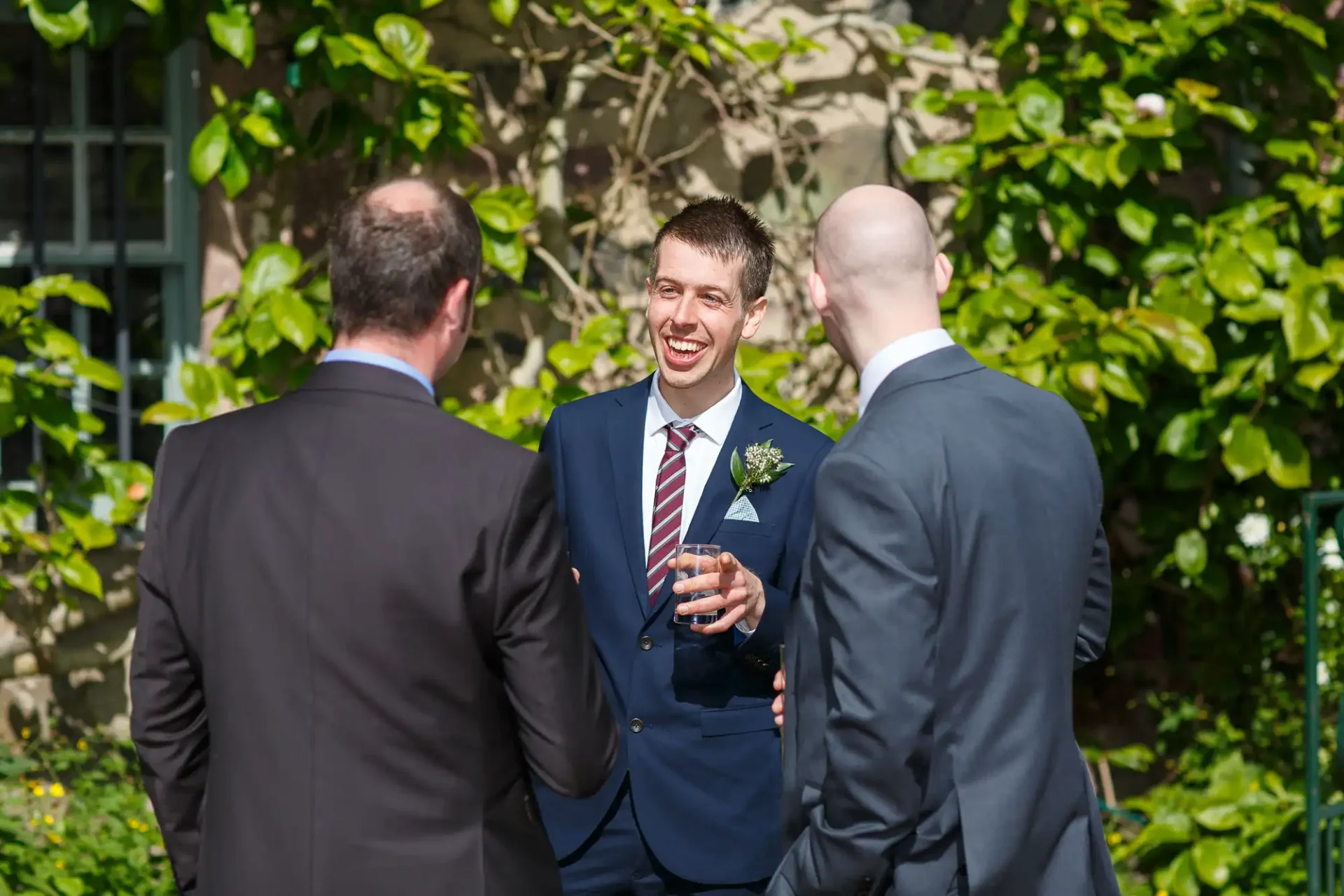 Man in a blue suit and striped tie laughing with two other men in suits at an outdoor gathering, holding a wine glass.