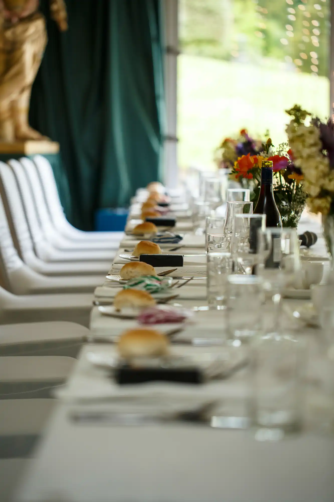 Elegant dining table set for an event with plates, glasses, and floral centerpieces, extending towards a window with natural light.