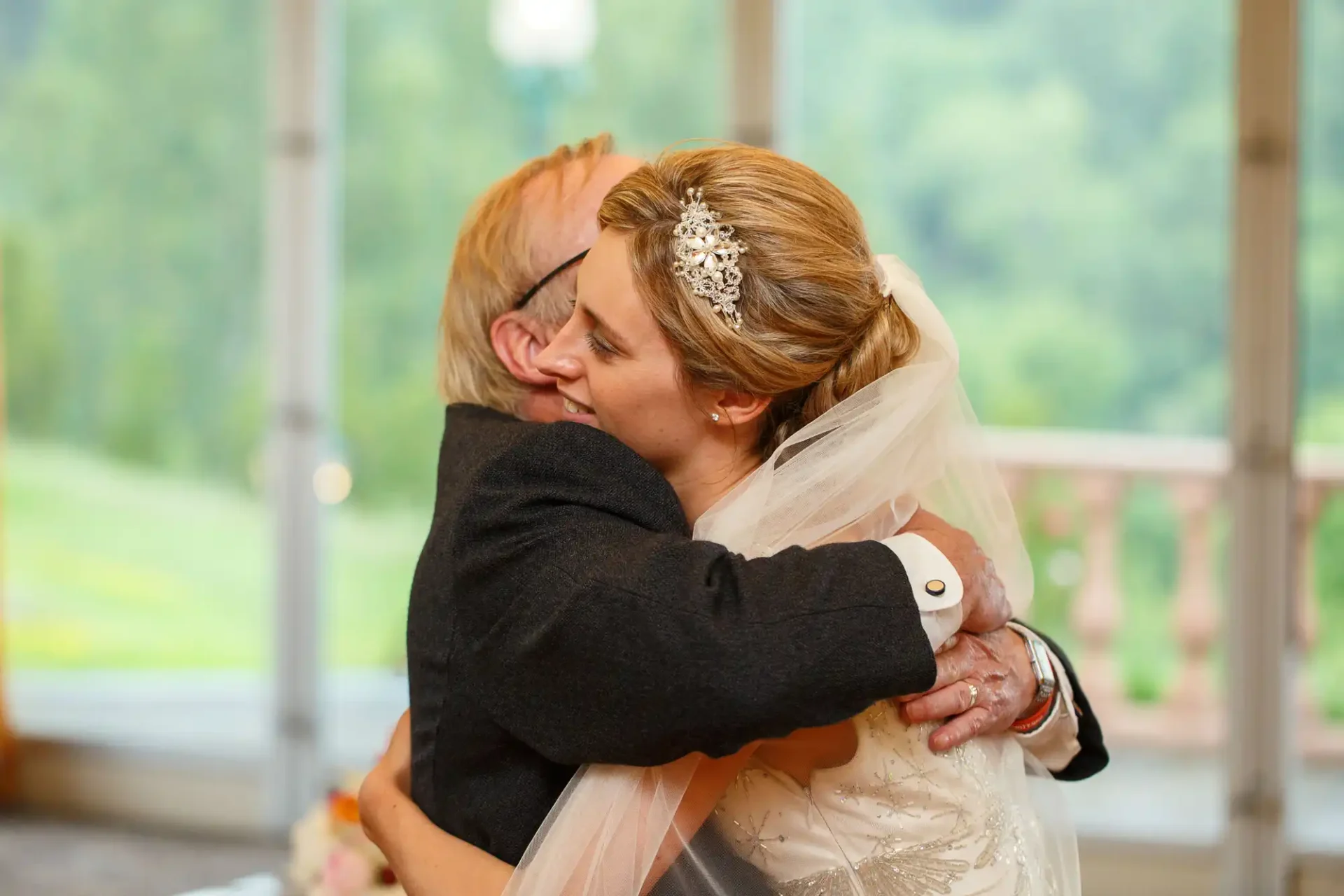 A bride in a white dress and tiara hugs an older man in a suit at her wedding, with a softly blurred green landscape visible through large windows behind them.