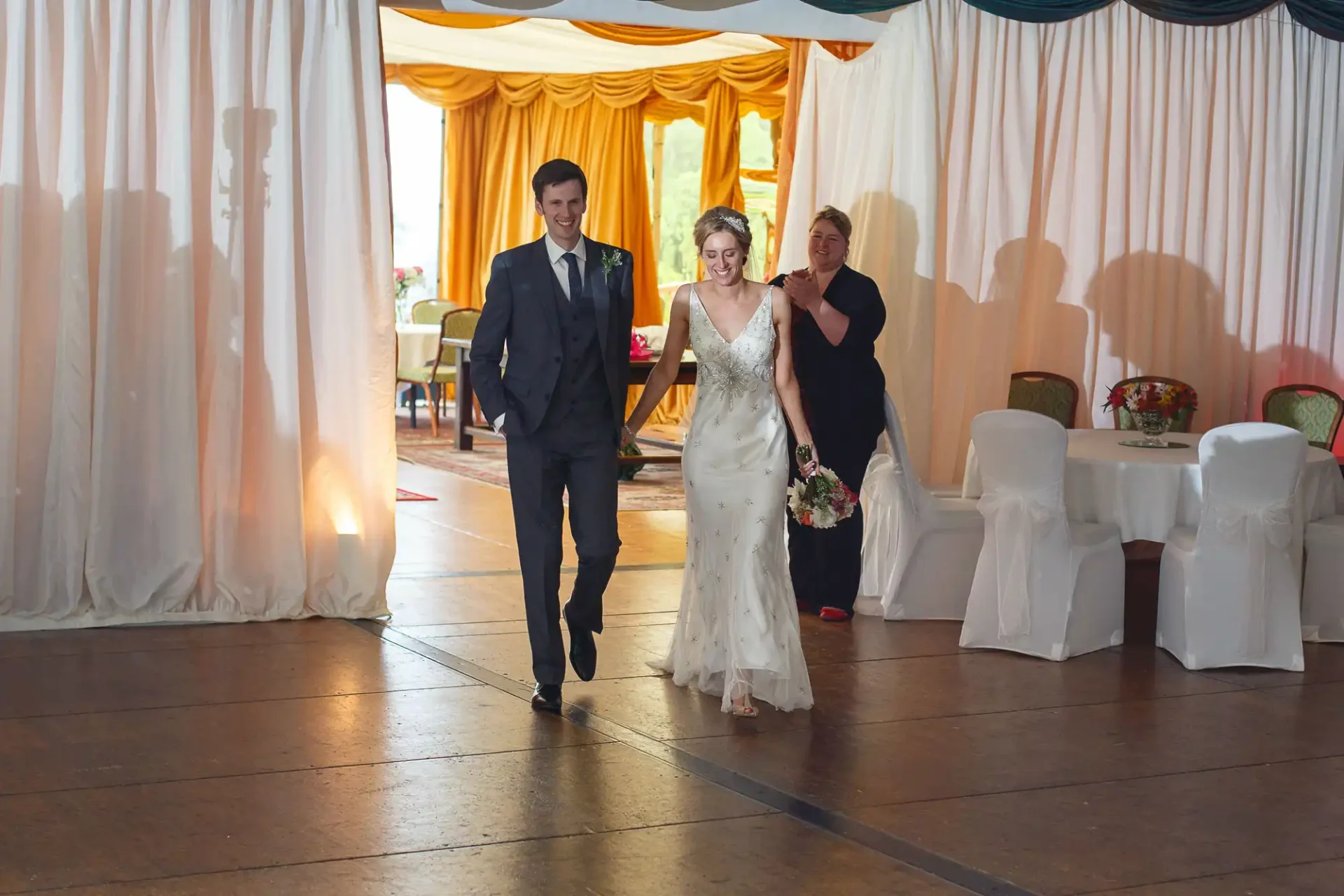 A bride and groom walk hand in hand into a reception hall, smiling broadly, as a woman adjusts the bride's veil from behind.