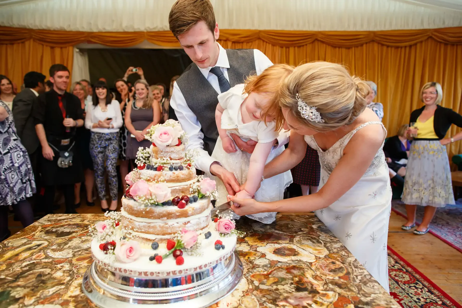 A bride and groom, holding a small child, cut their wedding cake surrounded by guests in a decorated tent.