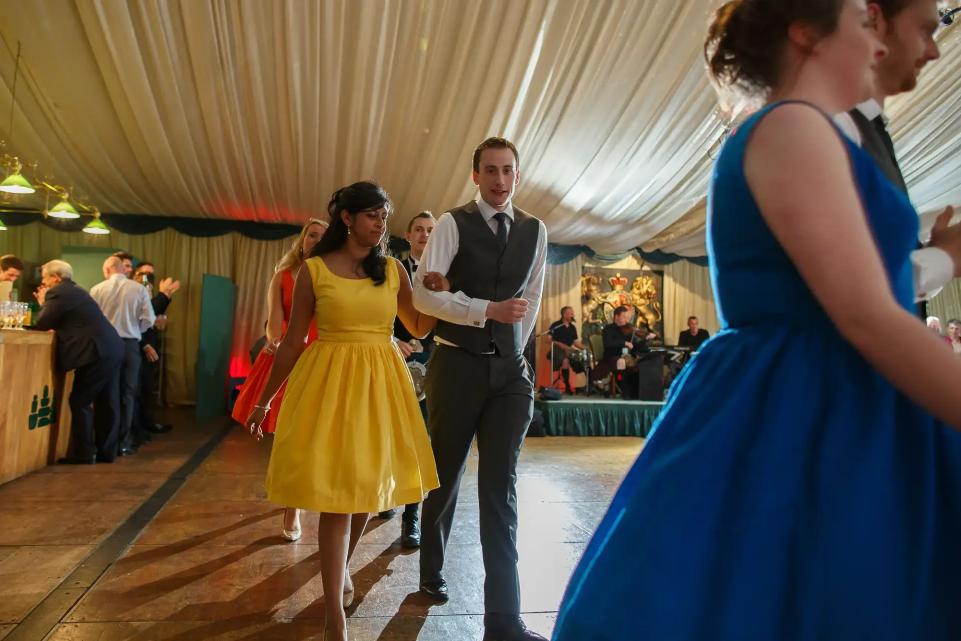 A man in a suit and a woman in a yellow dress walk hand in hand through a busy event tent, with other attendees in formal wear visible around them.