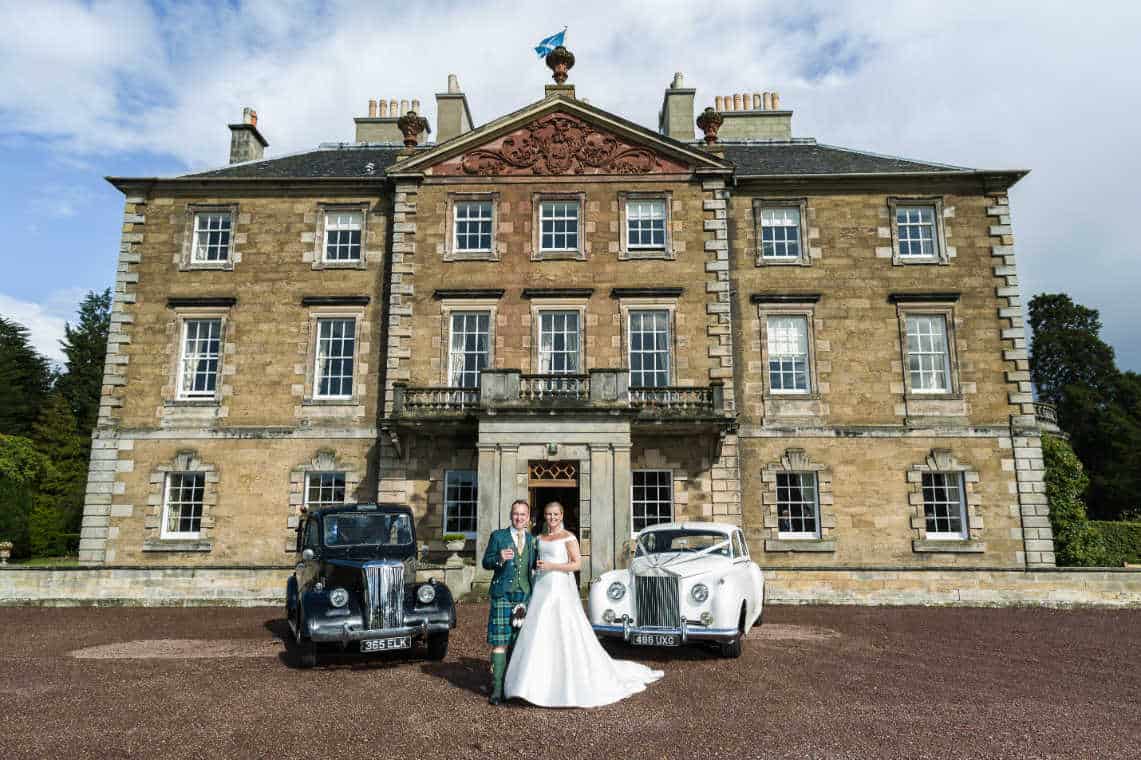 A newlywed couple stands in front of a grand historic mansion, Gilmerton House, flanked by two vintage cars. The groom wears a kilt, and the bride wears a white dress. The building is large, with many windows, stone walls, and a Scottish flag flying above the entrance.