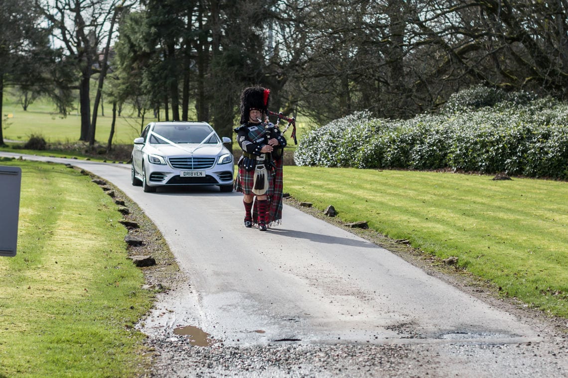 A bagpiper in traditional scottish attire walking beside a road, while a white car drives past on a sunny day.