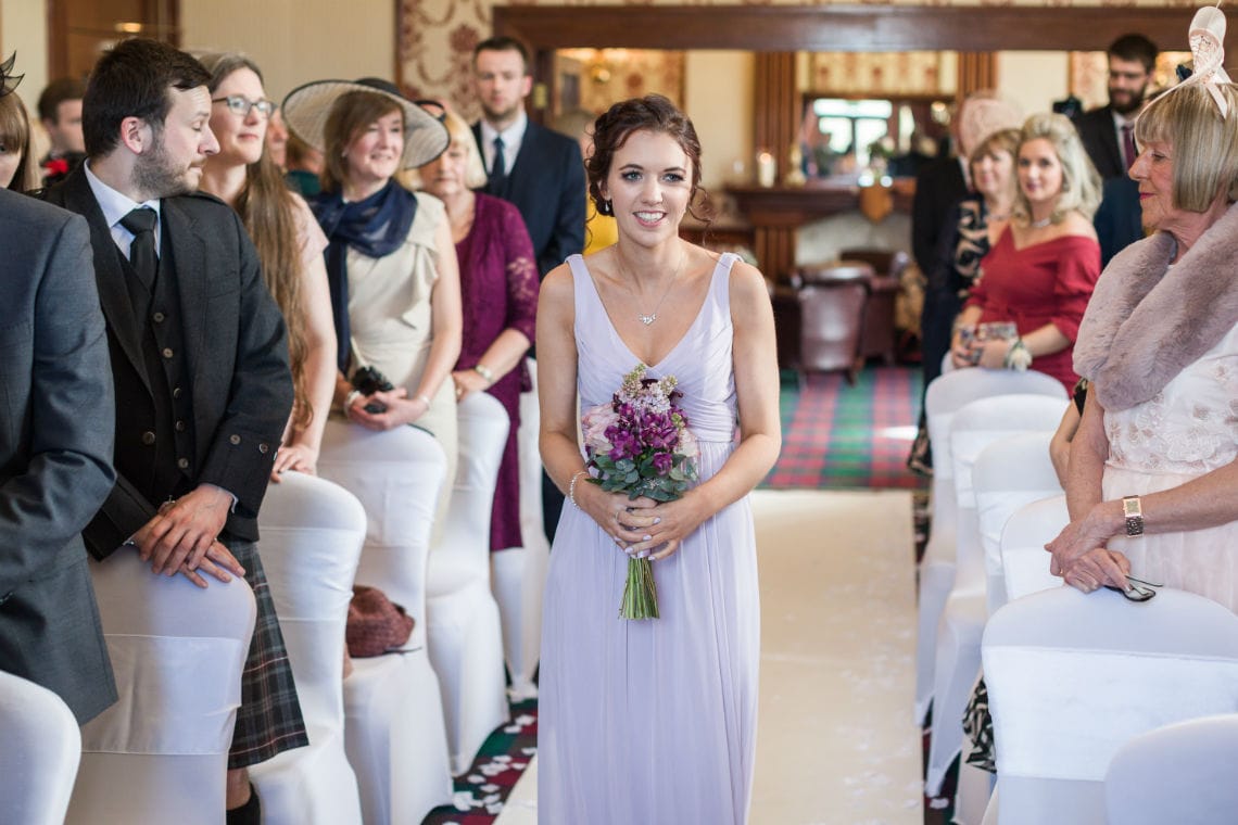 A bride walking down the aisle holding a bouquet, surrounded by guests in a well-lit room during a wedding ceremony.