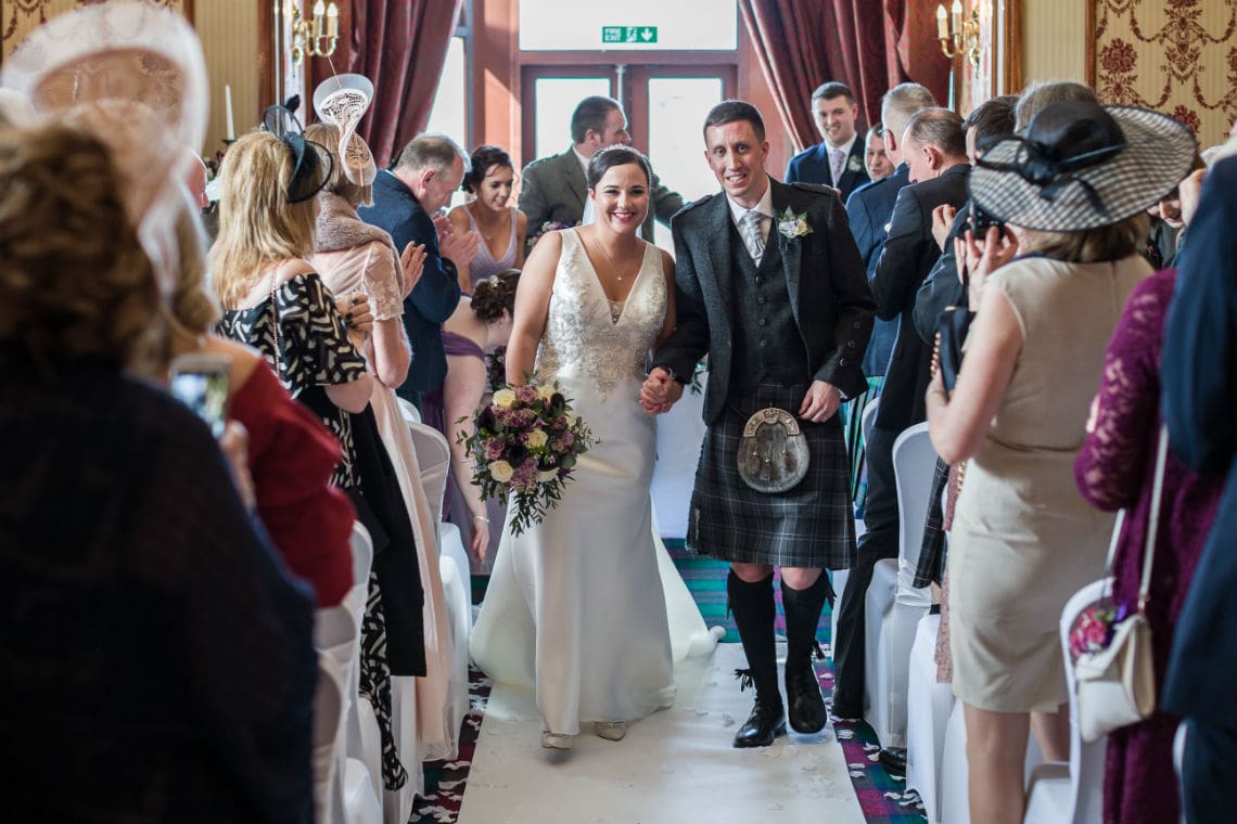 Bride and groom smiling as they walk through a crowd of guests, the groom wearing a kilt and the bride in a white dress, holding a bouquet.