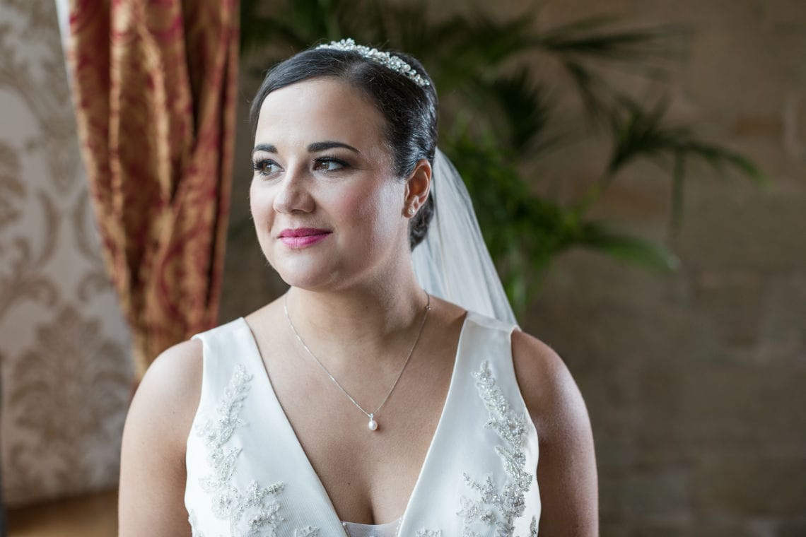 A bride in a white gown with lace detailing smiles softly, looking off to the side in a well-lit room with plants and draped curtains in the background.