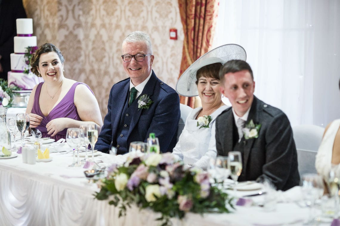 Four people at a wedding reception table, laughing and dressed formally, with floral centerpieces and elegant table settings.
