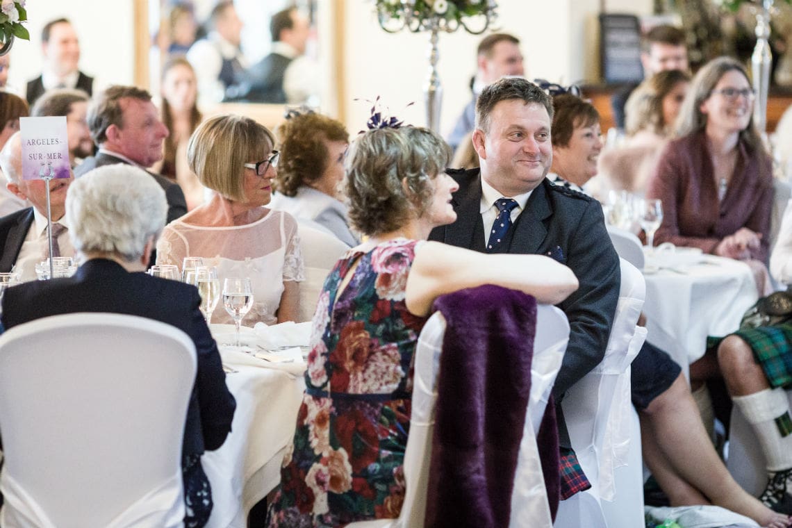 Guests seated at tables during a wedding reception, smiling and engaged in conversation. a man in a dark suit exchanges smiles with a woman in a floral dress.