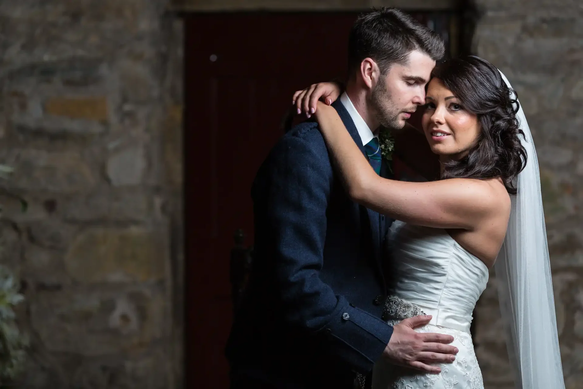 A bride and groom embrace lovingly, illuminated by soft light against a dark stone wall backdrop inside Glencorse Kirk.