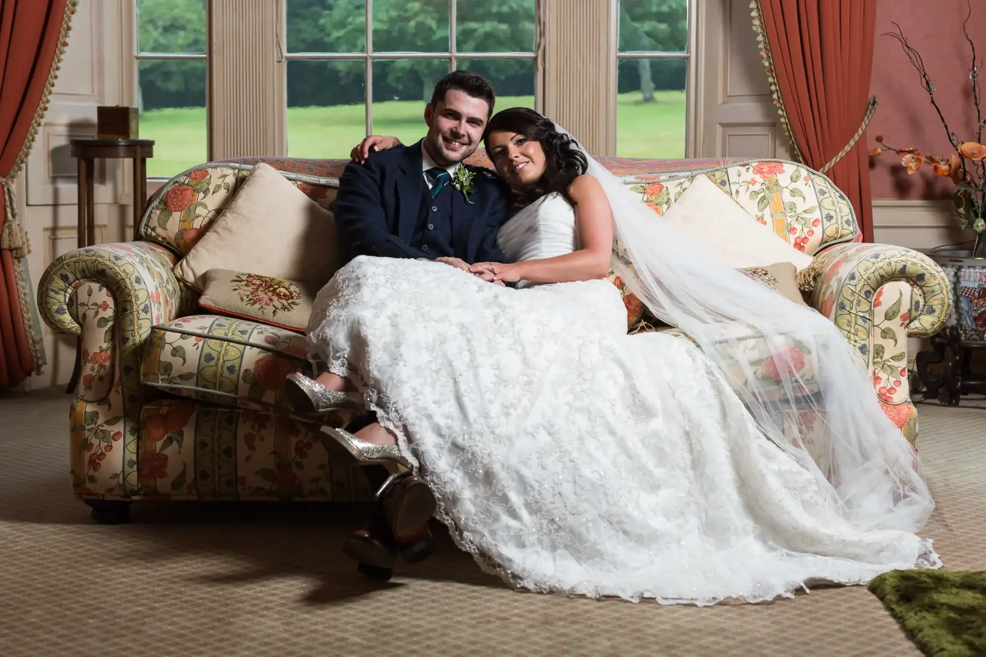 A newlywed couple smiling at Glencorse House, seated on a floral sofa in an elegant room, the bride in a white lace gown and the groom in a dark suit with a green boutonniere.
