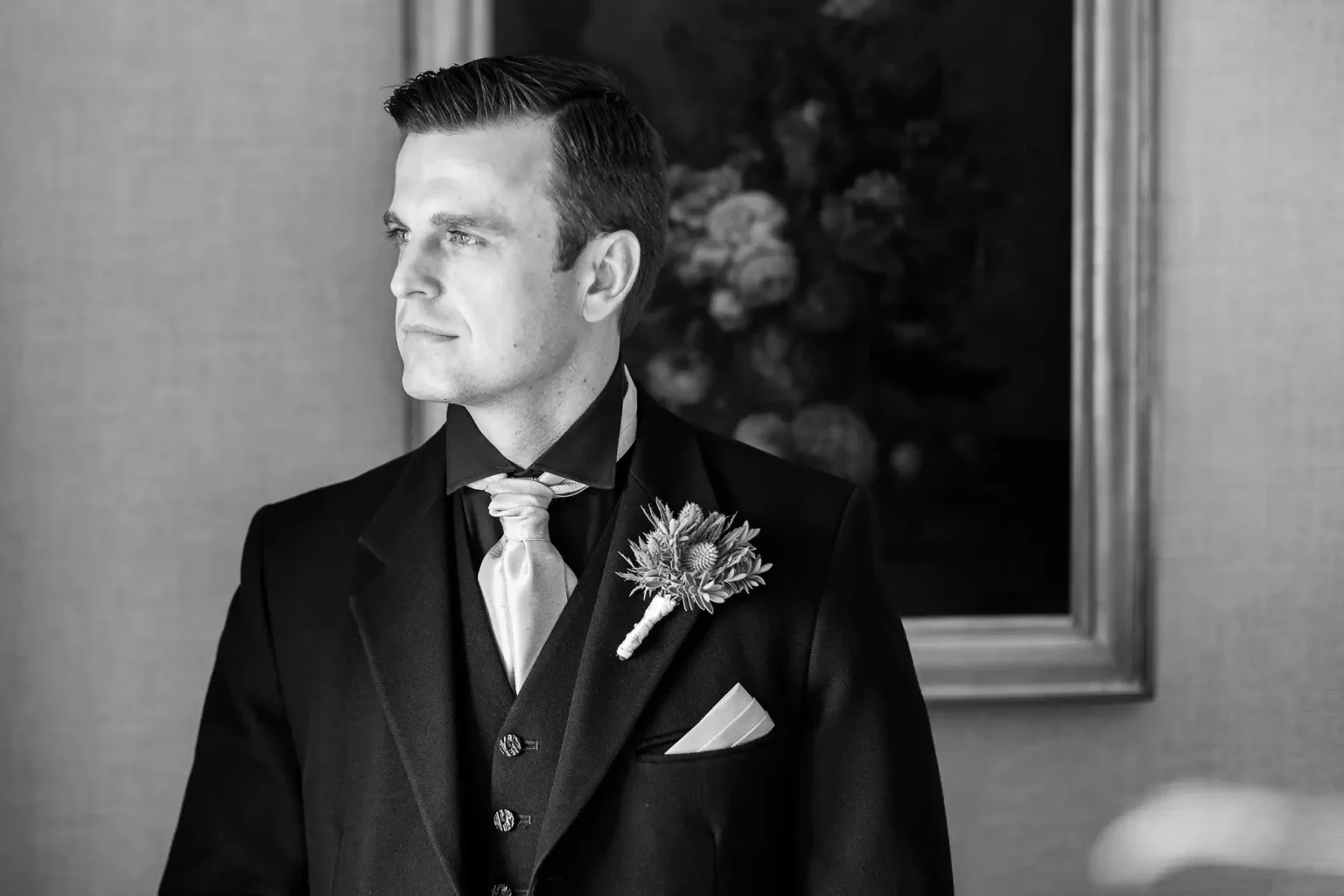 Black and white photography of a groom in a formal black suit, white shirt, and light-coloured cravat stands in front of a framed floral painting. He has neatly styled hair and is looking thoughtfully to the left. A buttonhole is pinned to his lapel. The image is black and white.