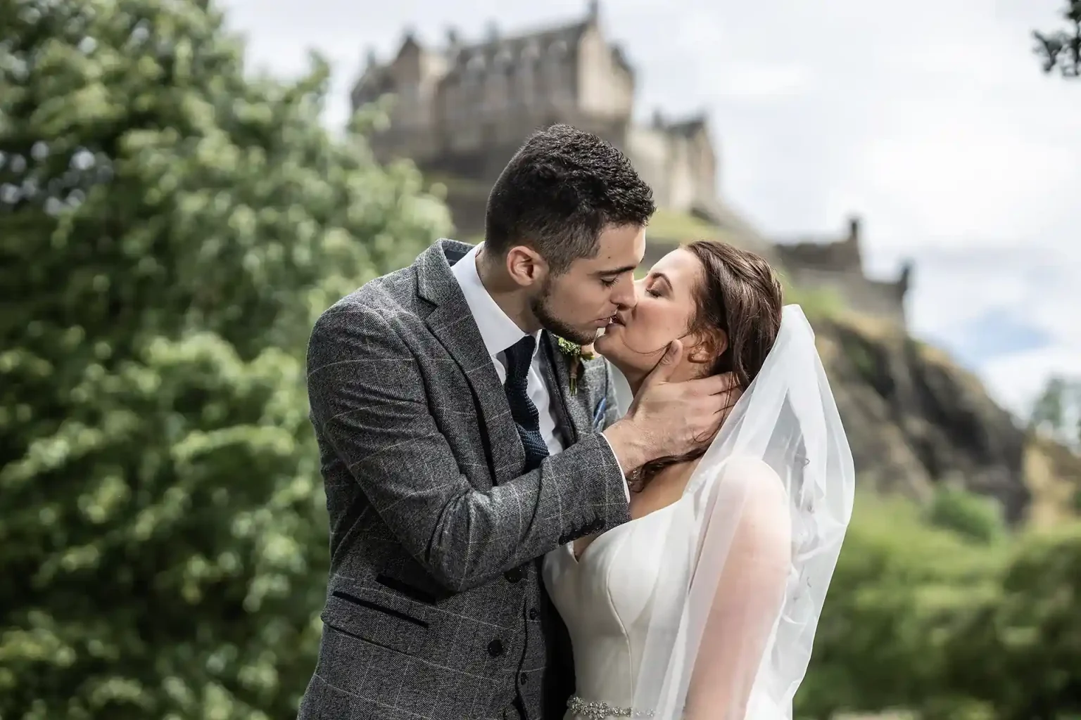 A bride and groom share a kiss outdoors with a castle in the background. The groom wears a gray suit and the bride wears a white dress with a veil.