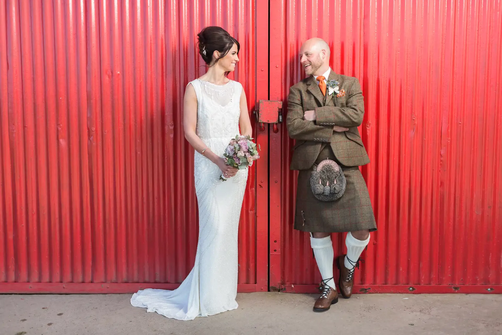 A bride in a white dress and a groom in a kilt sharing a cheerful moment against a vibrant red background at Kinkell Byre.