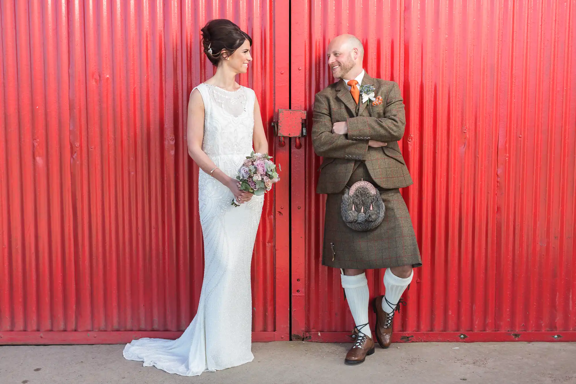 A bride in a white dress holding a bouquet stands smiling at a groom in a brown Scottish kilt outfit. They lean against a large red corrugated metal door, looking at each other affectionately. The groom’s arms are folded, and both appear happy and relaxed during their Kinkell Byre wedding photography session.