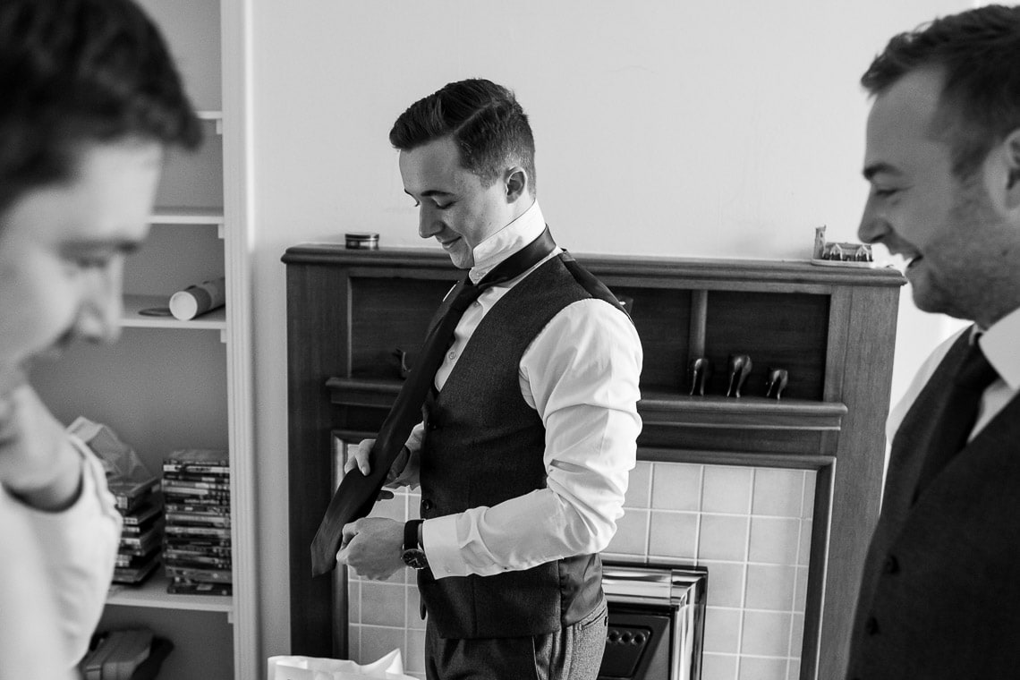 Man in vest and tie smiles while adjusting his sleeve, flanked by two other men in a room with a bookshelf.