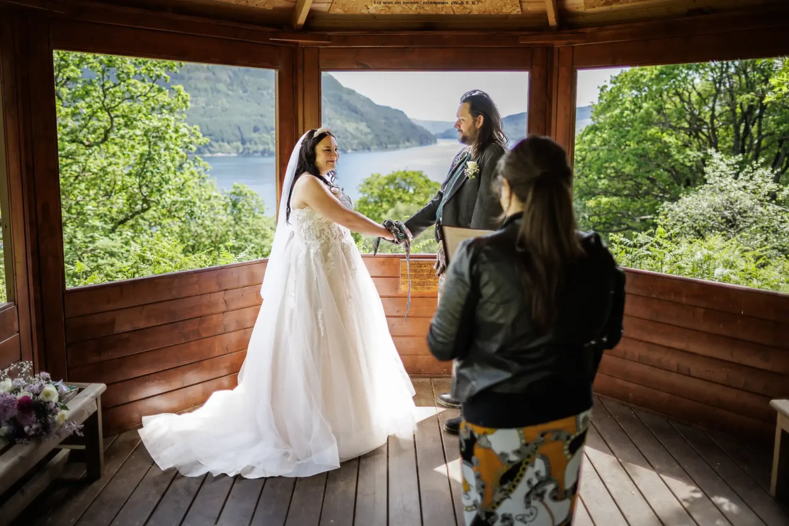 A bride and groom hold hands during a wedding ceremony in a wooden gazebo with a scenic forest and lake view, as an officiant stands nearby.
