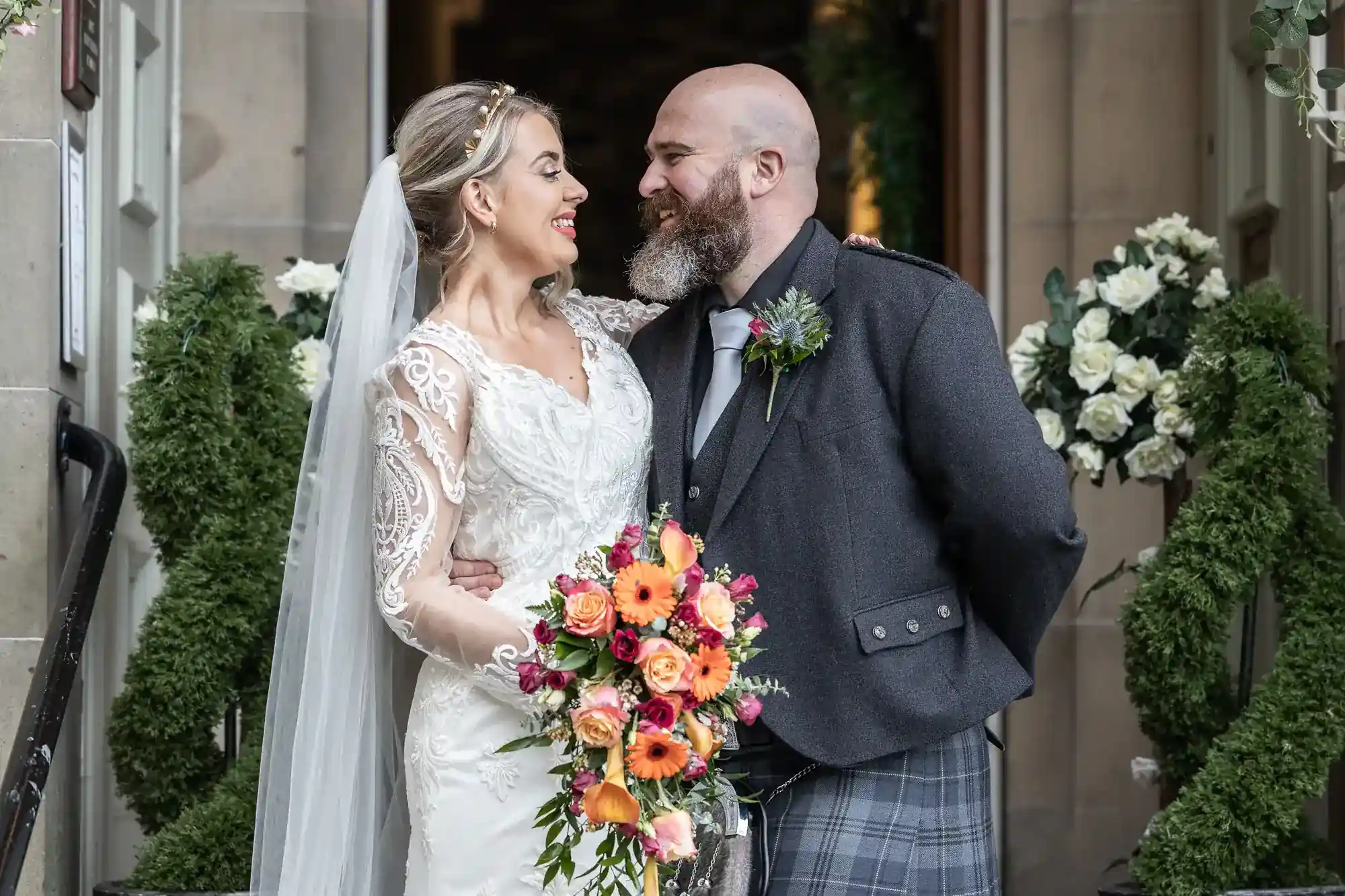 A couple, dressed in wedding attire, stand smiling at each other outside a building. The bride holds a colourful bouquet, and greenery with white flowers can be seen in the background at Mansfield House Hotel.