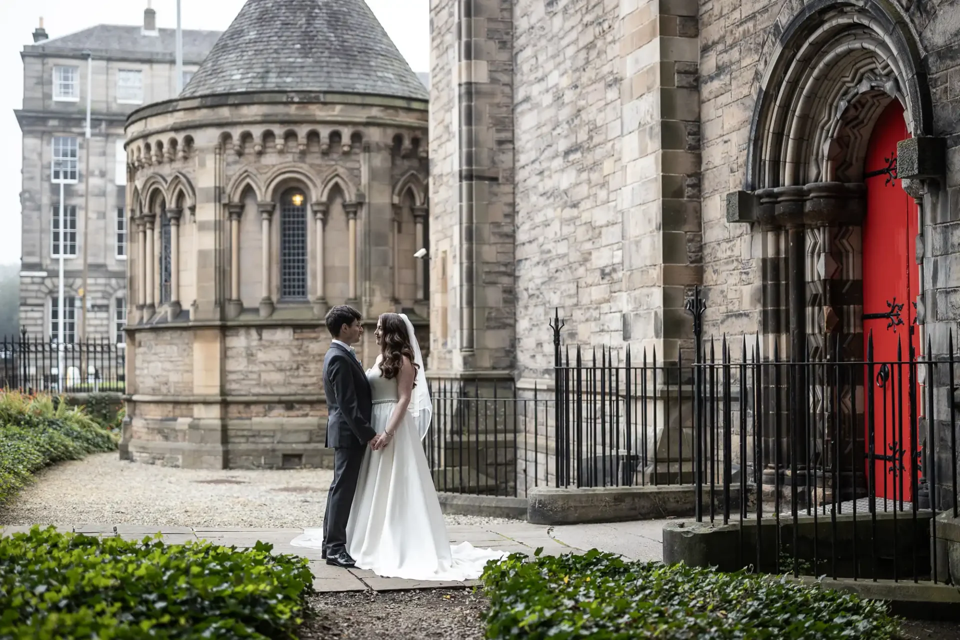 A bride and groom stand facing each other outside in the The Private Garden, at Mansfield Traquair, a historic stone church, with arched windows and a red door .