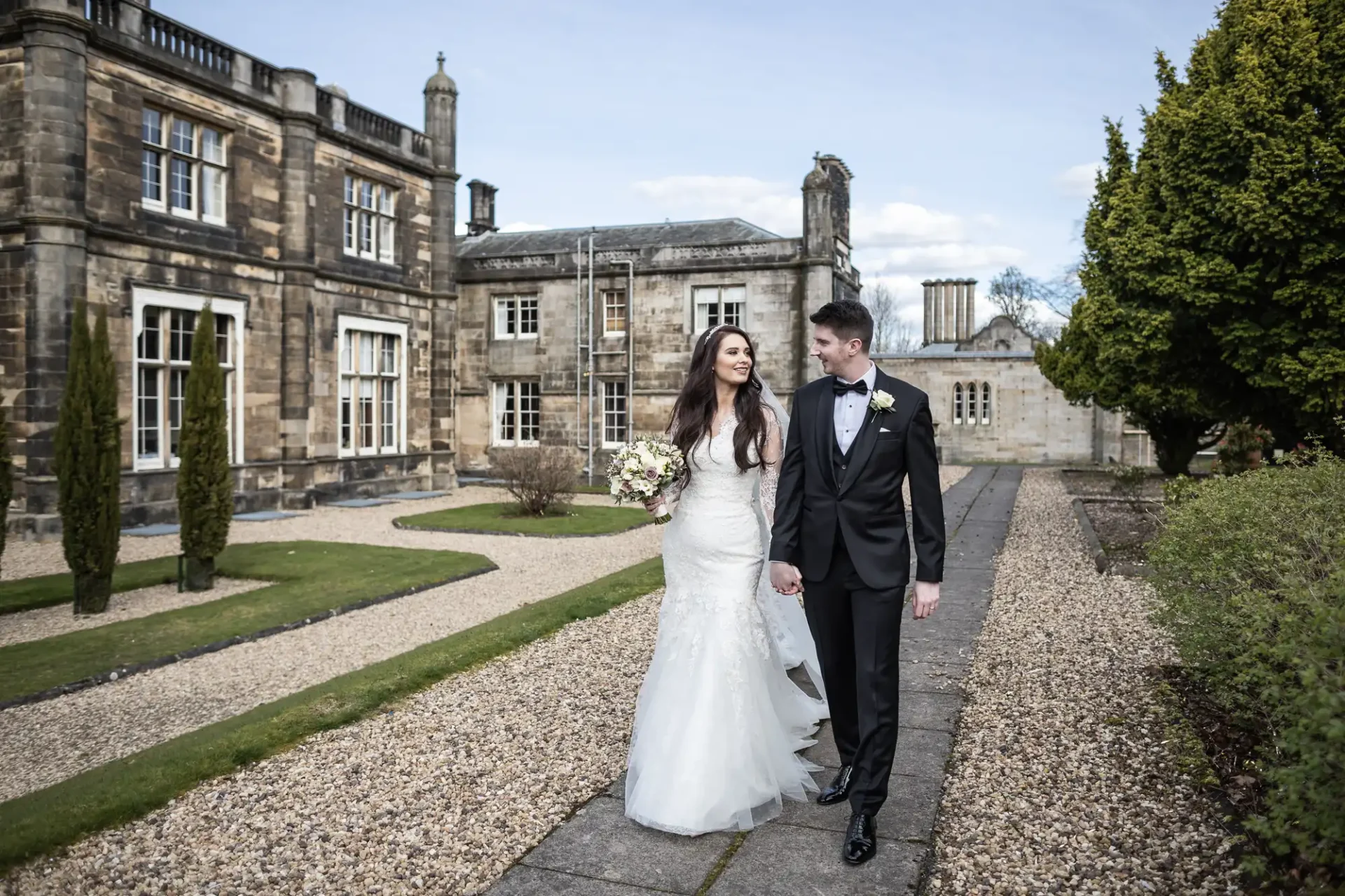 Mar Hall wedding photographer image of a couple in wedding attire holding hands, walking on a path in front of a historic stone building and manicured garden.