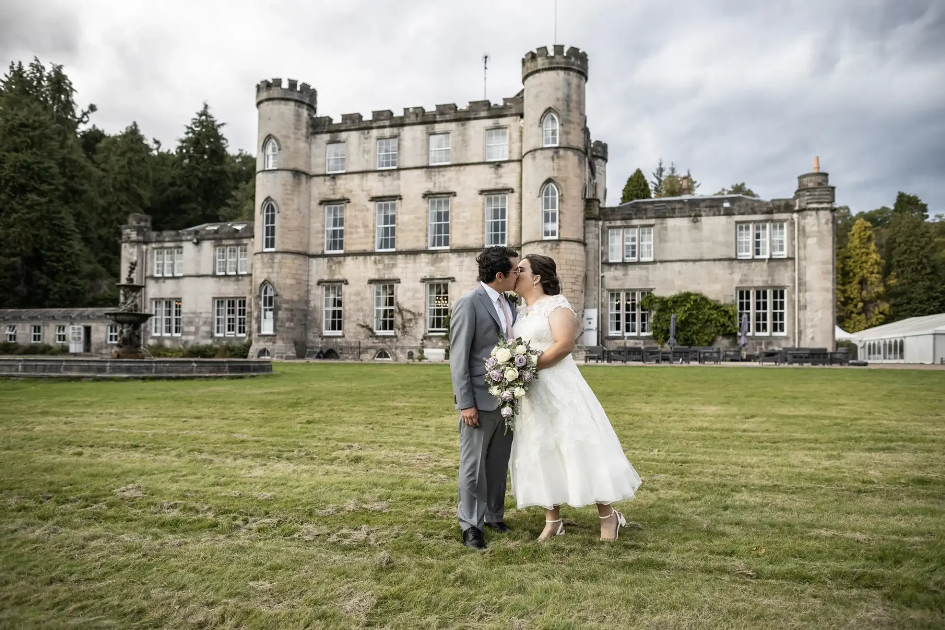 A couple dressed in wedding attire stand close together in front of Melville Castle, near Edinburgh, on a cloudy day. The bride holds a bouquet of flowers.