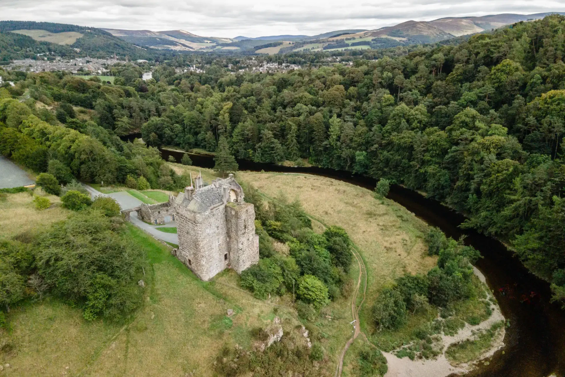 Aerial view of a partially ruined stone castle, Neidpath Castle, surrounded by lush greenery, with a river flowing nearby and hills in the background.