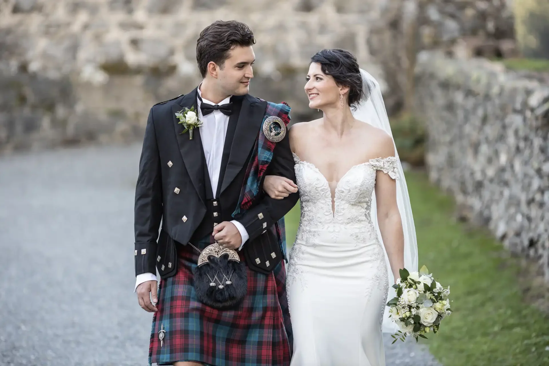 Neidpath Castle wedding photographer photo of a  bride and groom walking arm in arm outdoors. The groom wears a traditional Scottish kilt, and the bride wears a white off-the-shoulder wedding dress, holding a bouquet. They both are looking at each other.