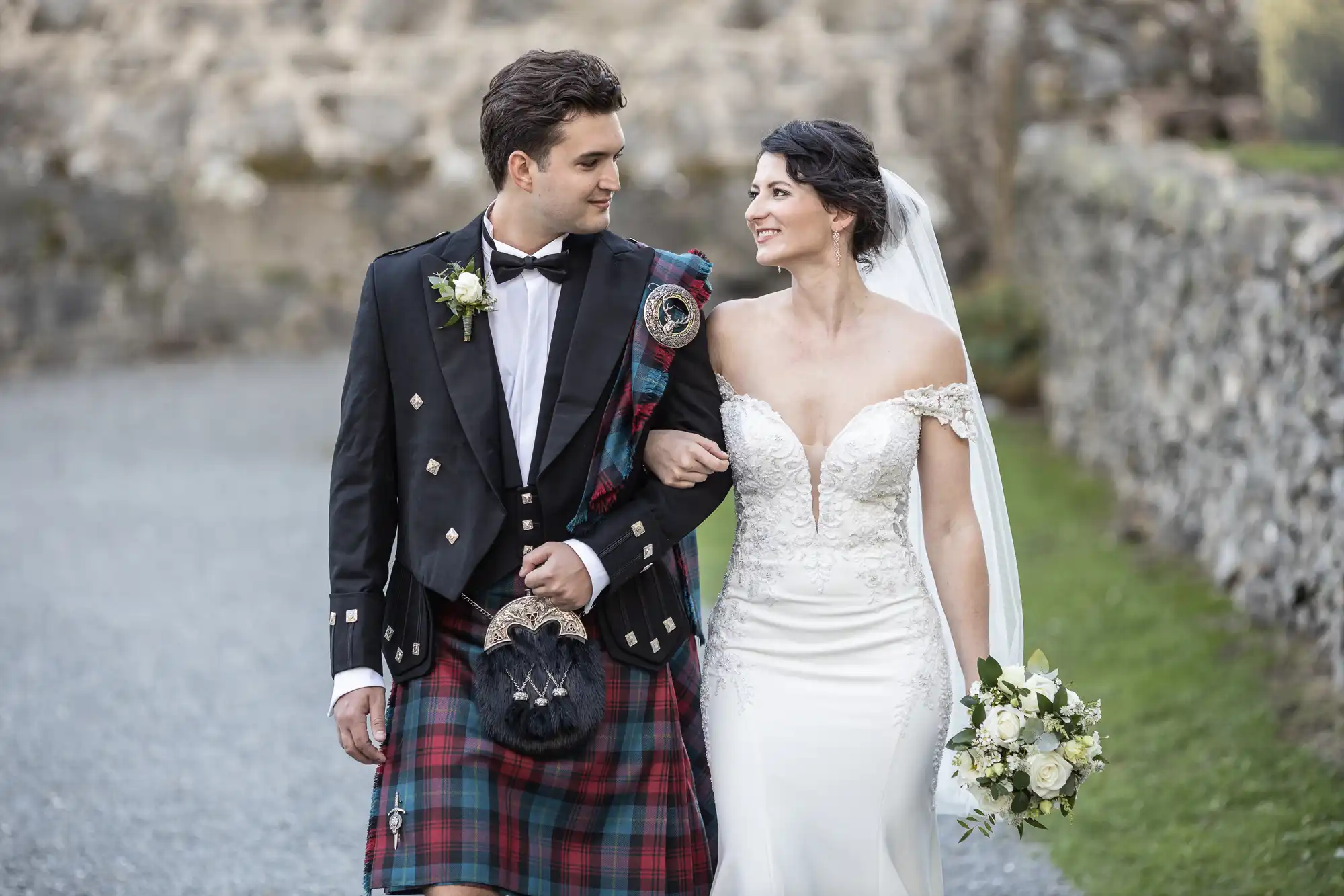 Neidpath Castle wedding photographer photo of a bride and groom walking arm in arm outdoors. The groom wears a traditional Scottish kilt, and the bride wears a white off-the-shoulder wedding dress, holding a bouquet. They both are looking at each other.
