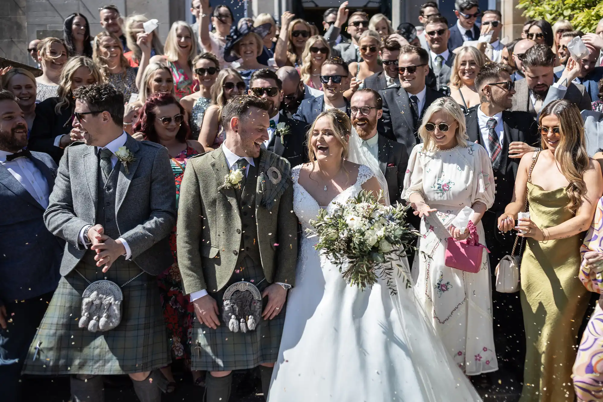 A bride and groom in wedding attire stand smiling among a large group of well-dressed guests during an outdoor celebration. Some guests are wearing sunglasses, and flower petals are in the air at Netherbyres House.