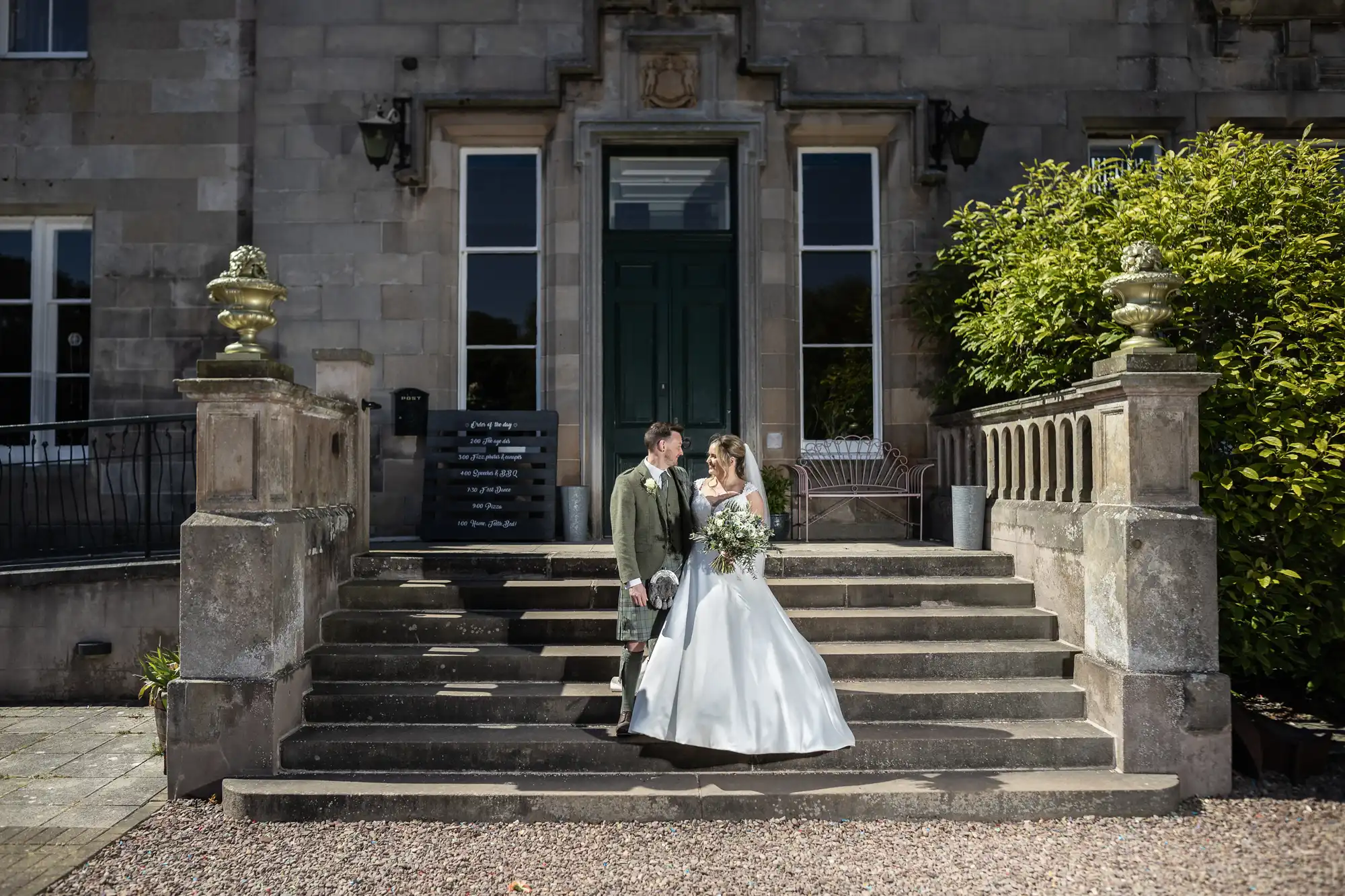 A newlywed couple stands on stone steps in front of a large building, the groom in a kilt and the bride in a white gown, holding a bouquet.