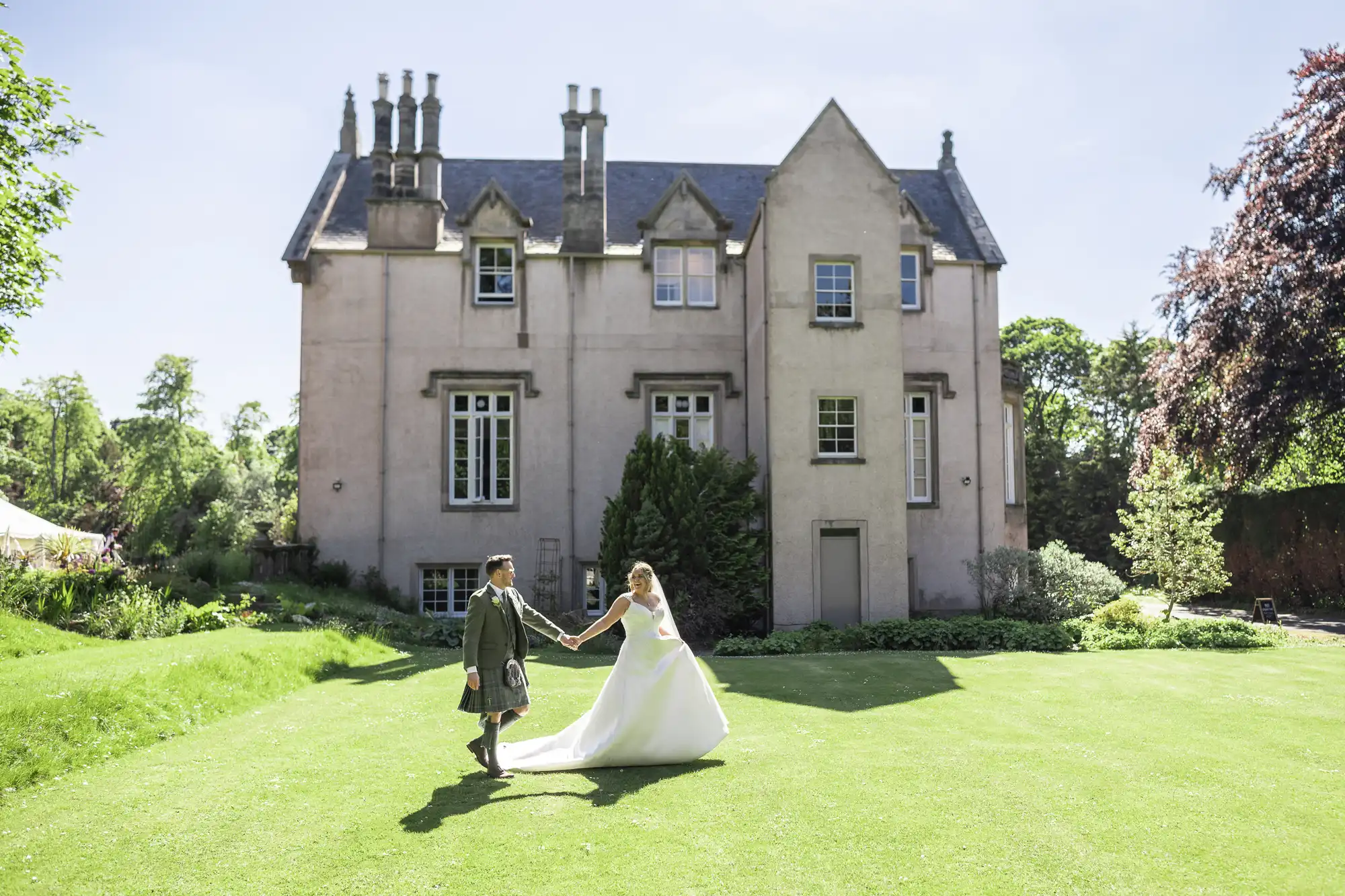 A couple, with the groom in a kilt and the bride in a white gown, hold hands on a green lawn in front of a large historic building, Netherbyres House.