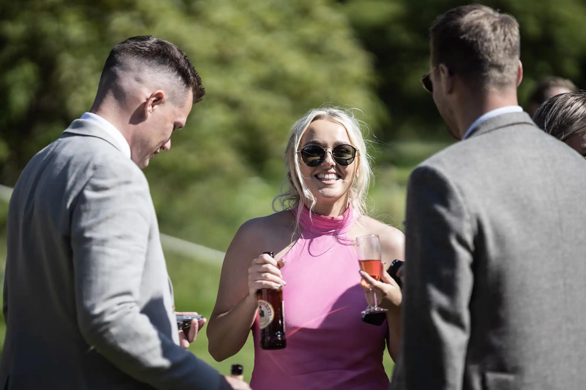 Three people talking outdoors, with one woman in a pink dress holding a drink and smiling. Two men in grey suits face her, also holding drinks. Trees are in the background.