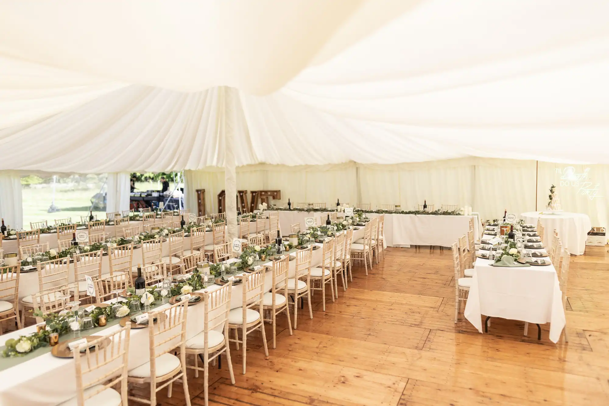A wedding reception setup inside a white, fabric-draped marquee tent featuring long wooden tables lined with white chairs and simple floral centerpieces.